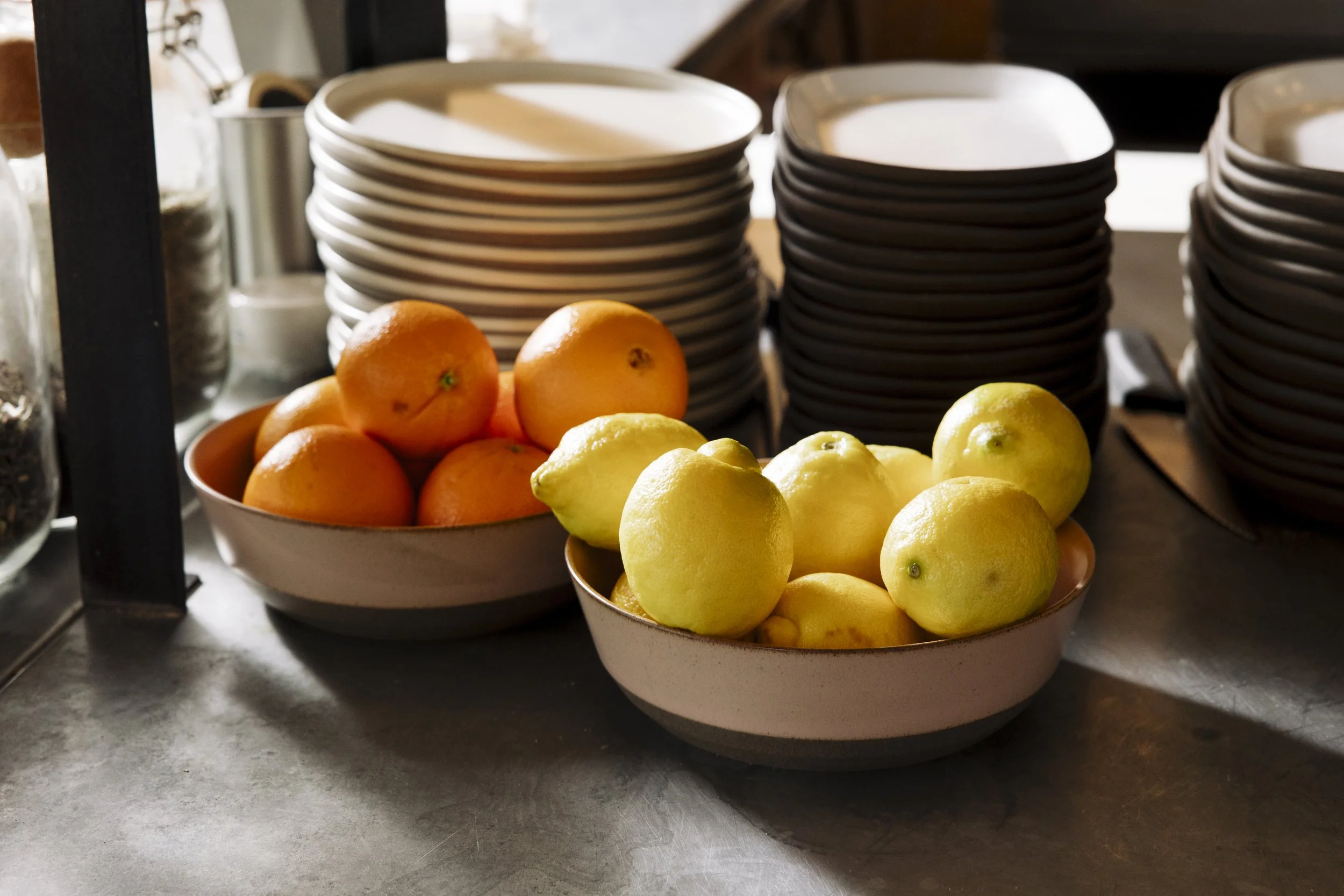 Bowl of oranges and lemons on a dark countertop with stacks of plates in the background.