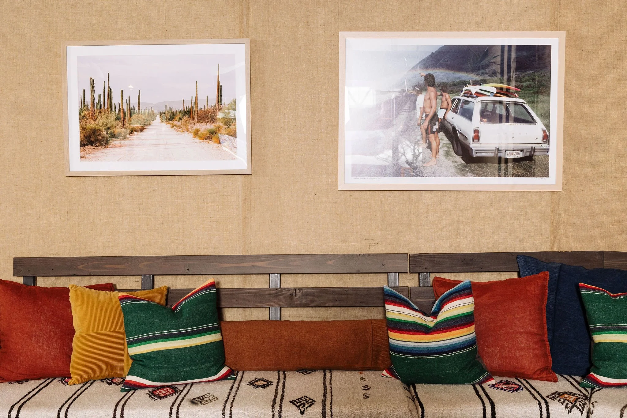 A collection of colorful striped pillows arranged on a wooden bench against a beige wall with two framed photographs hanging above.