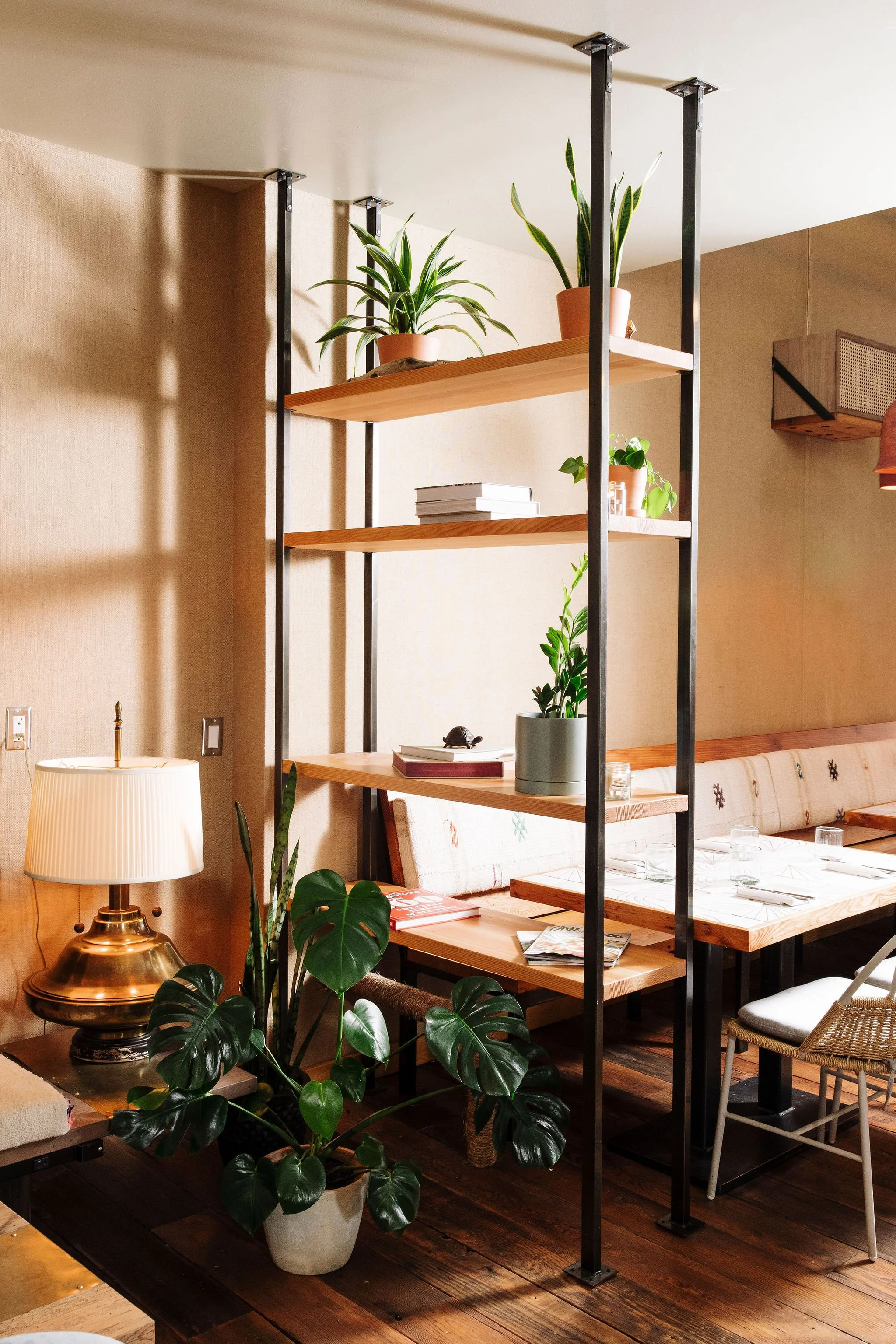 Interior of a cozy restaurant with wooden tables, glassware, potted plants, a lamp, and a bookshelf, emphasizing a warm and inviting atmosphere.
