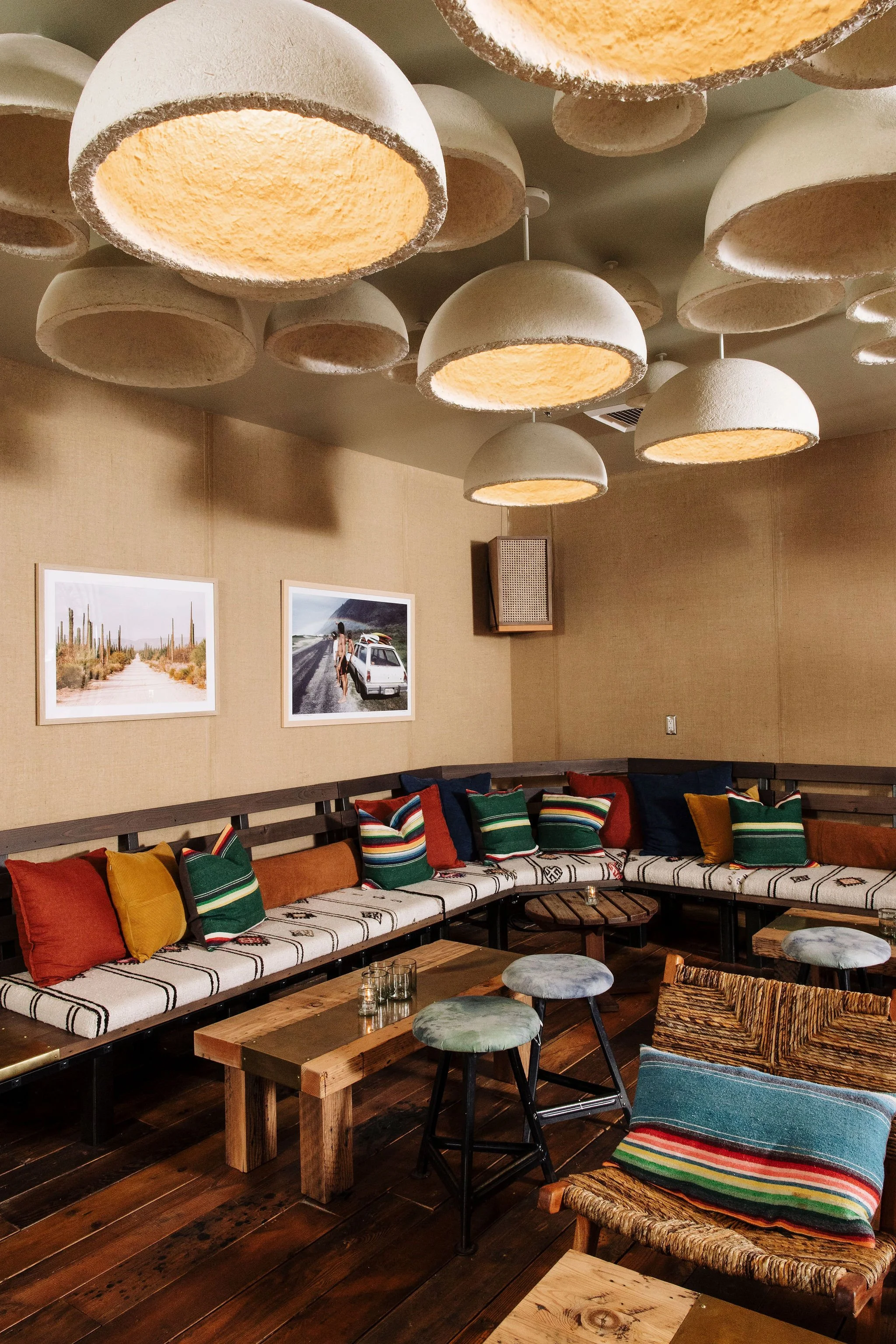 Interior of a cozy cafe or lounge area with a long cushioned bench lined with colorful pillows. Statement pendant lights with textured shades hang from the ceiling. Two framed landscape photos are on the wall, and a wooden table with small candles is