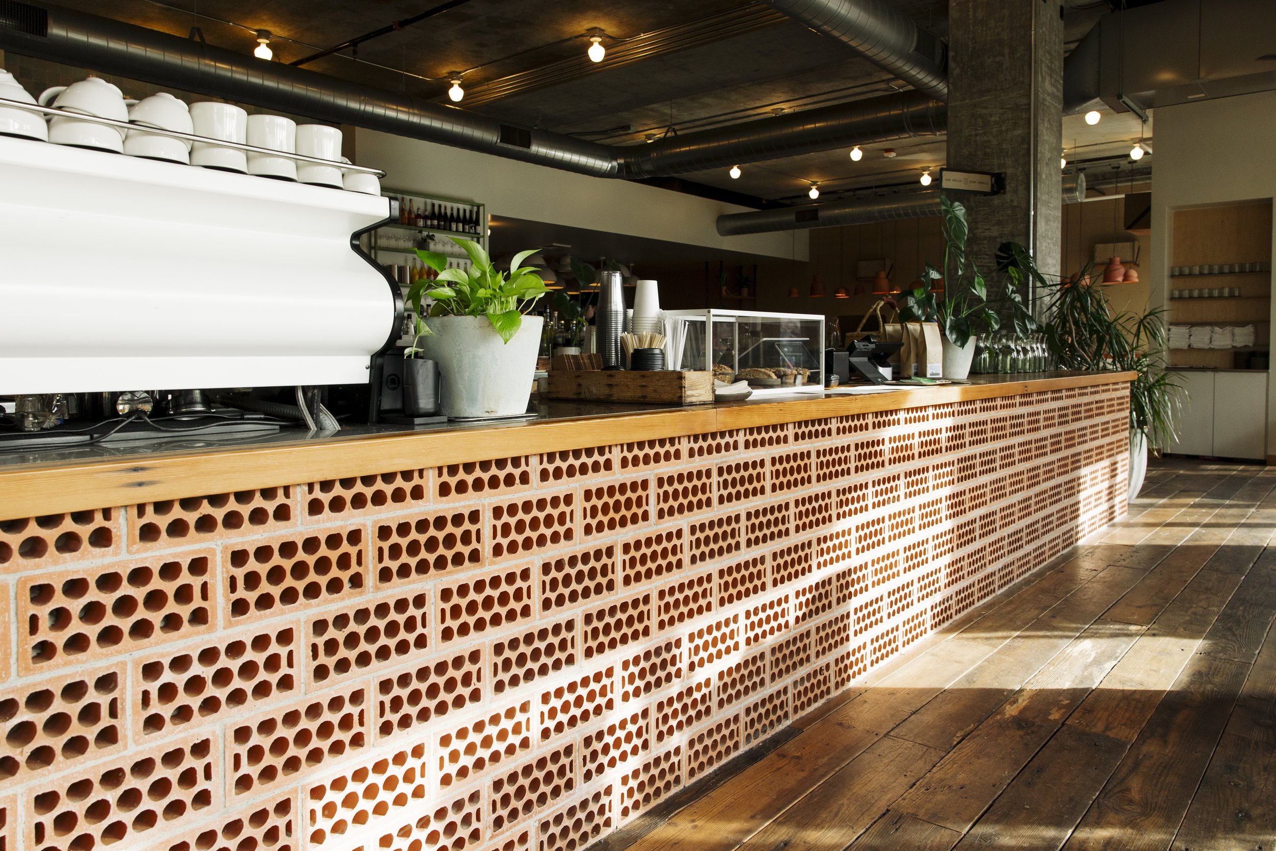 Interior of a modern cafe or restaurant with a wooden counter that has a decorative brick pattern. The counter has potted plants, disposable coffee cups, and a display case with baked goods. The ceiling features exposed ductwork, and there is warm li