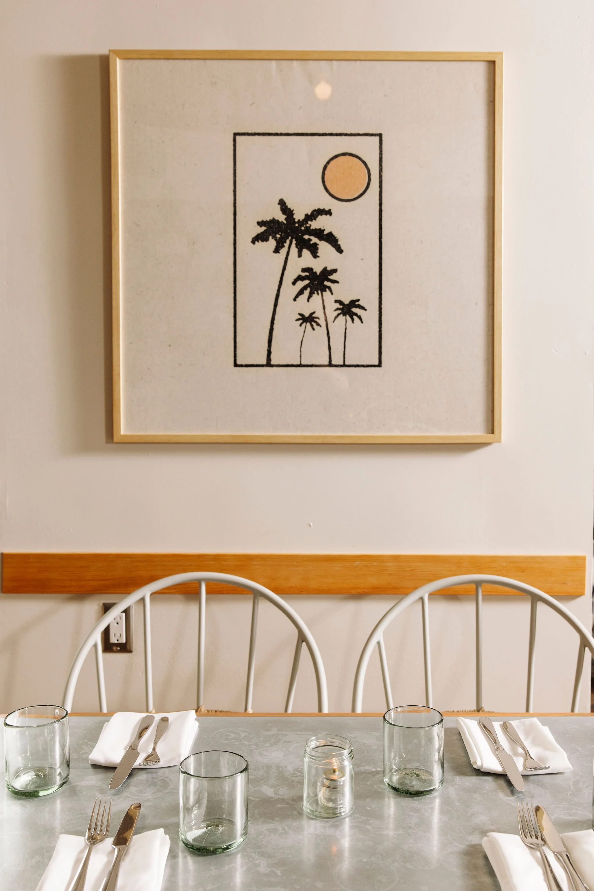 A table set for dining with four glasses and silverware on white napkins, in front of a wall illuminated by soft lighting. Framed beach-themed artwork with palm trees and a sun hangs on the wall.