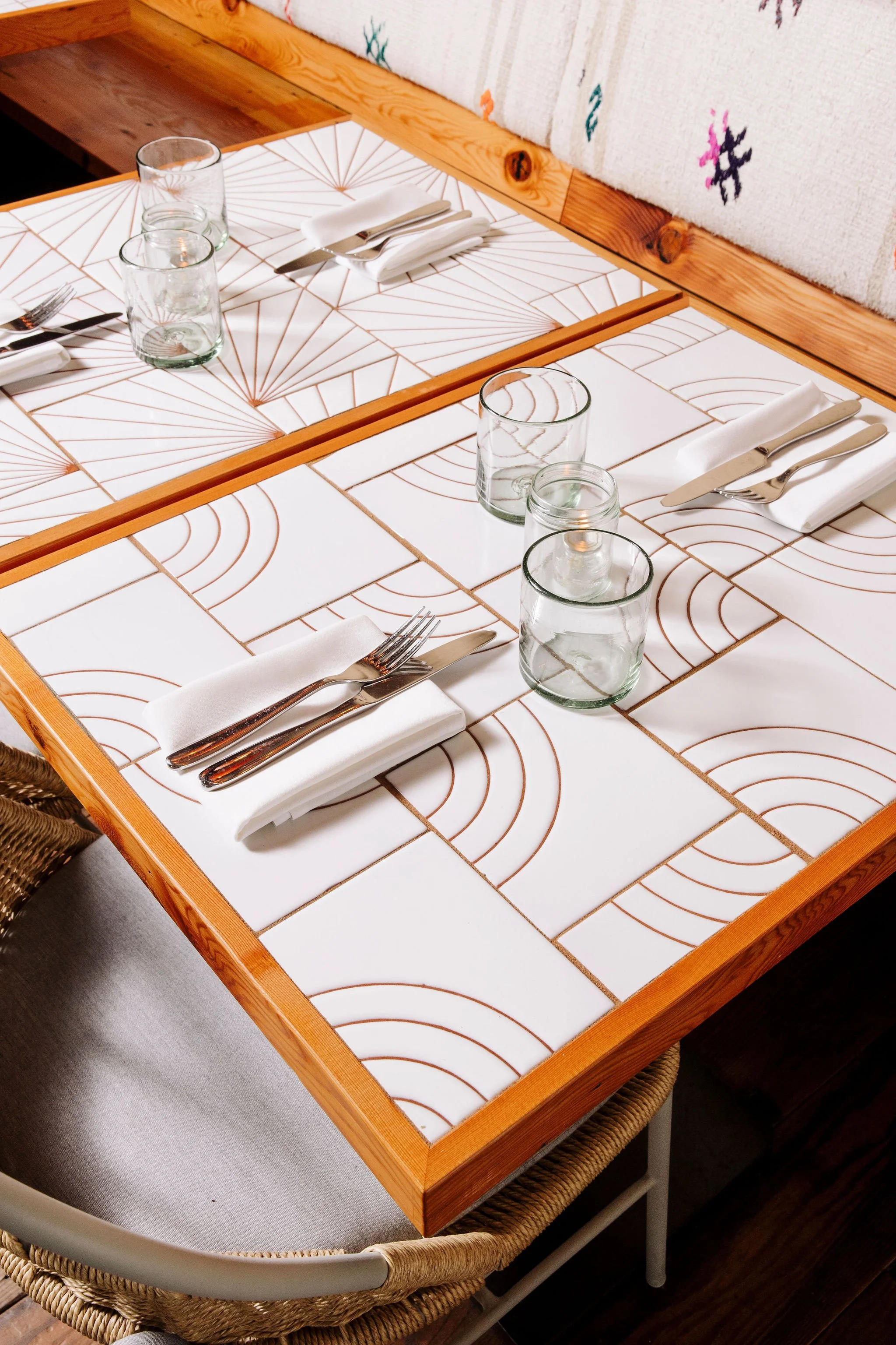 A dining table set with white tiles featuring geometric brown lines, white cloth napkins, silverware, clear water glasses, and small jars, with a wicker chair and part of a white tablecloth underneath.