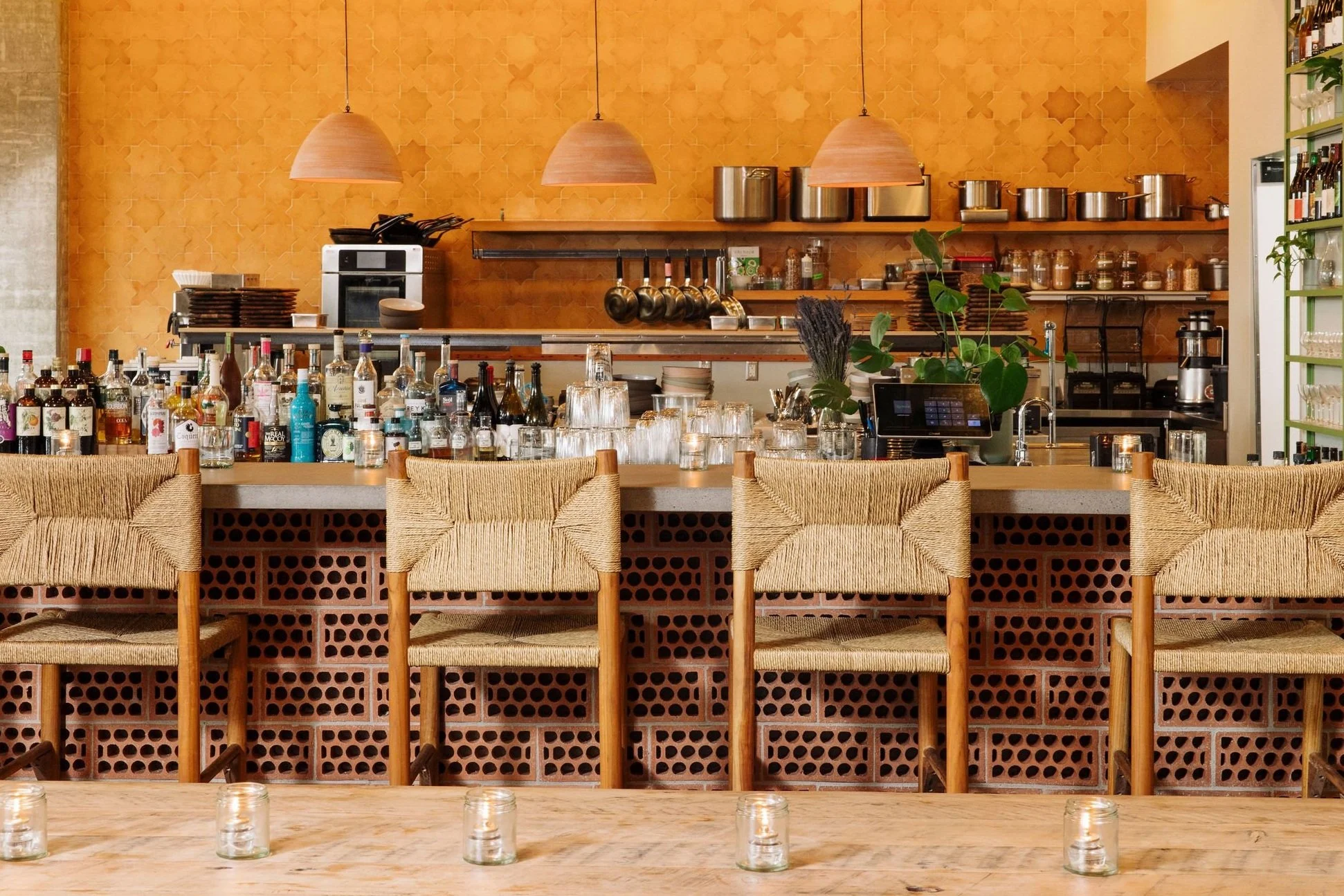 A restaurant bar with a counter, bottles, glasses, a plant, and hanging light fixtures.