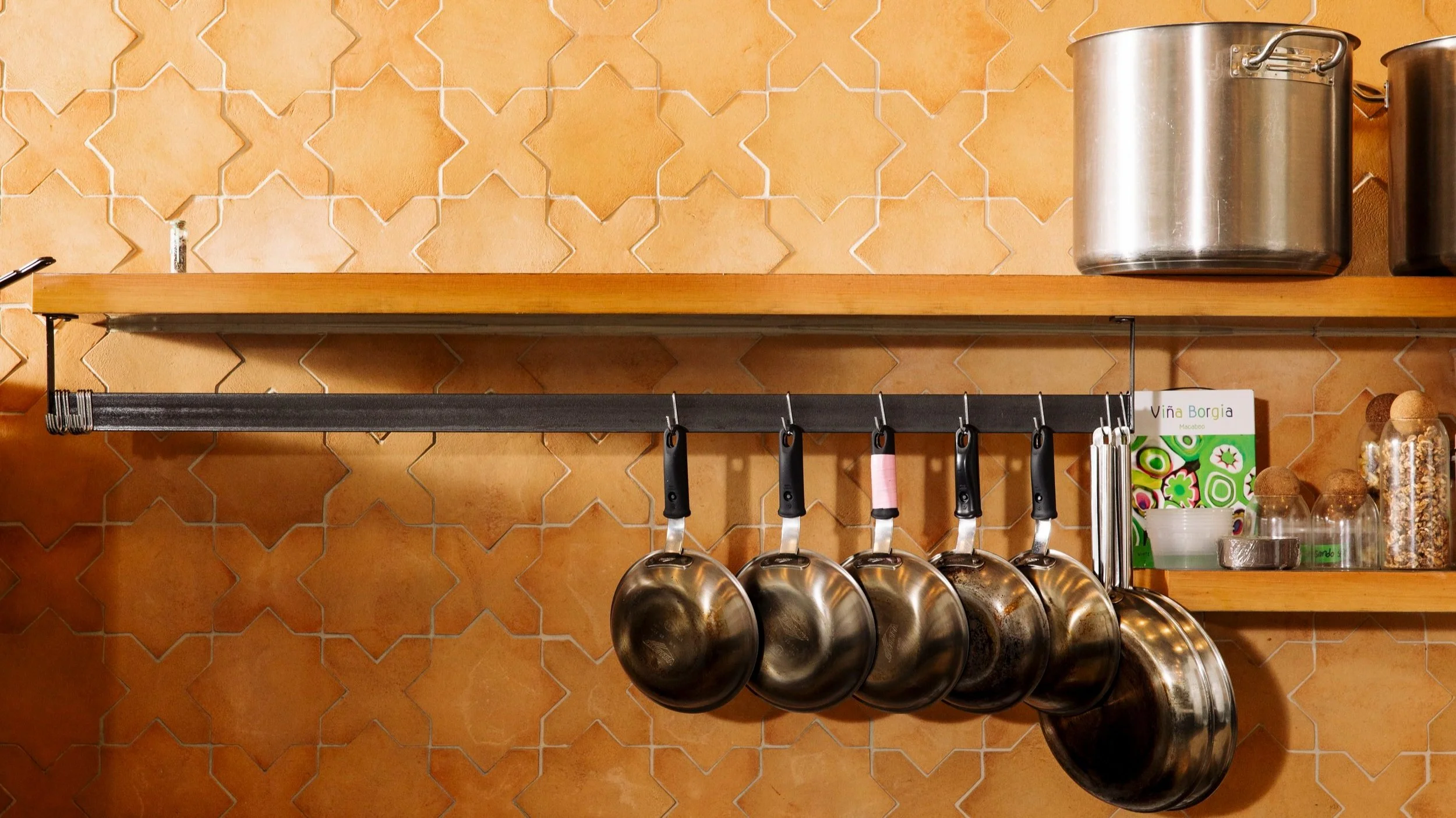 Kitchen wall with hanging pots, pans, and kitchen utensils on a wooden shelf against a decorative tiled wall.