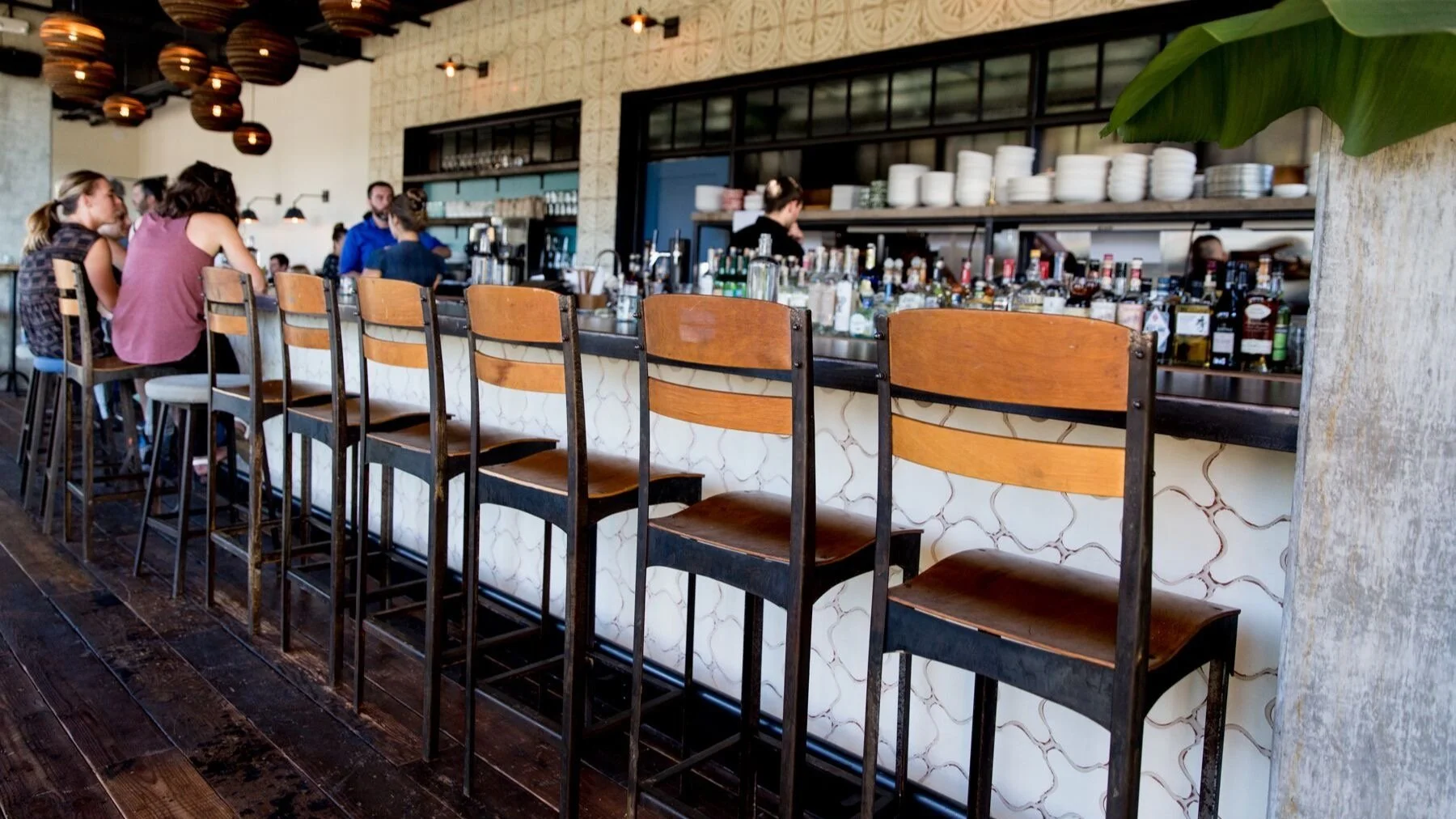 Inside a modern bar with a long counter and high wooden chairs, some patrons sitting at the bar, and bartenders preparing drinks, decorated with pendant lights and a large green leaf in the foreground.