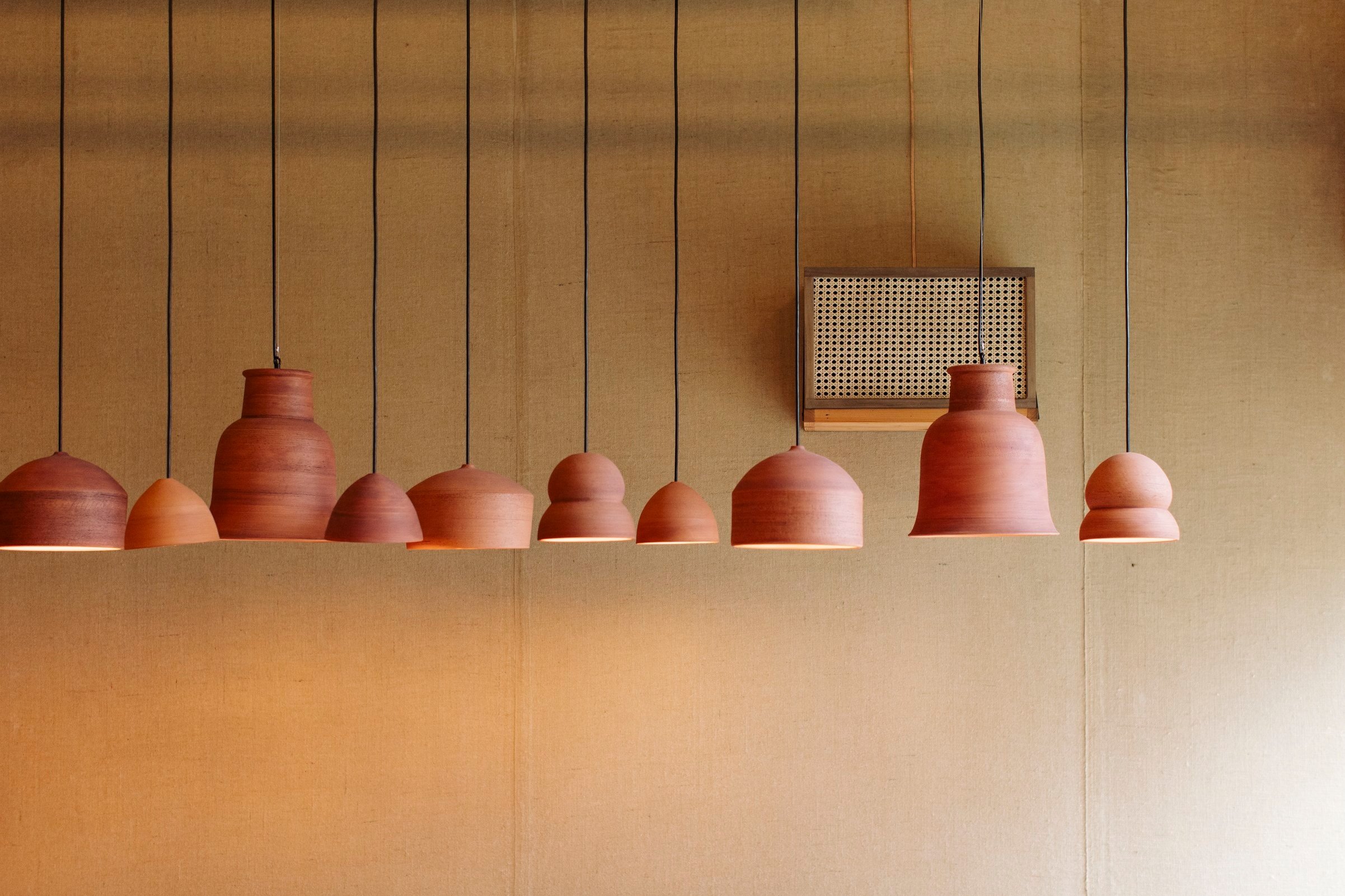 A ceiling with a row of terracotta-colored pendant lights hanging from black cords and a square wooden vent cover on a beige wall.