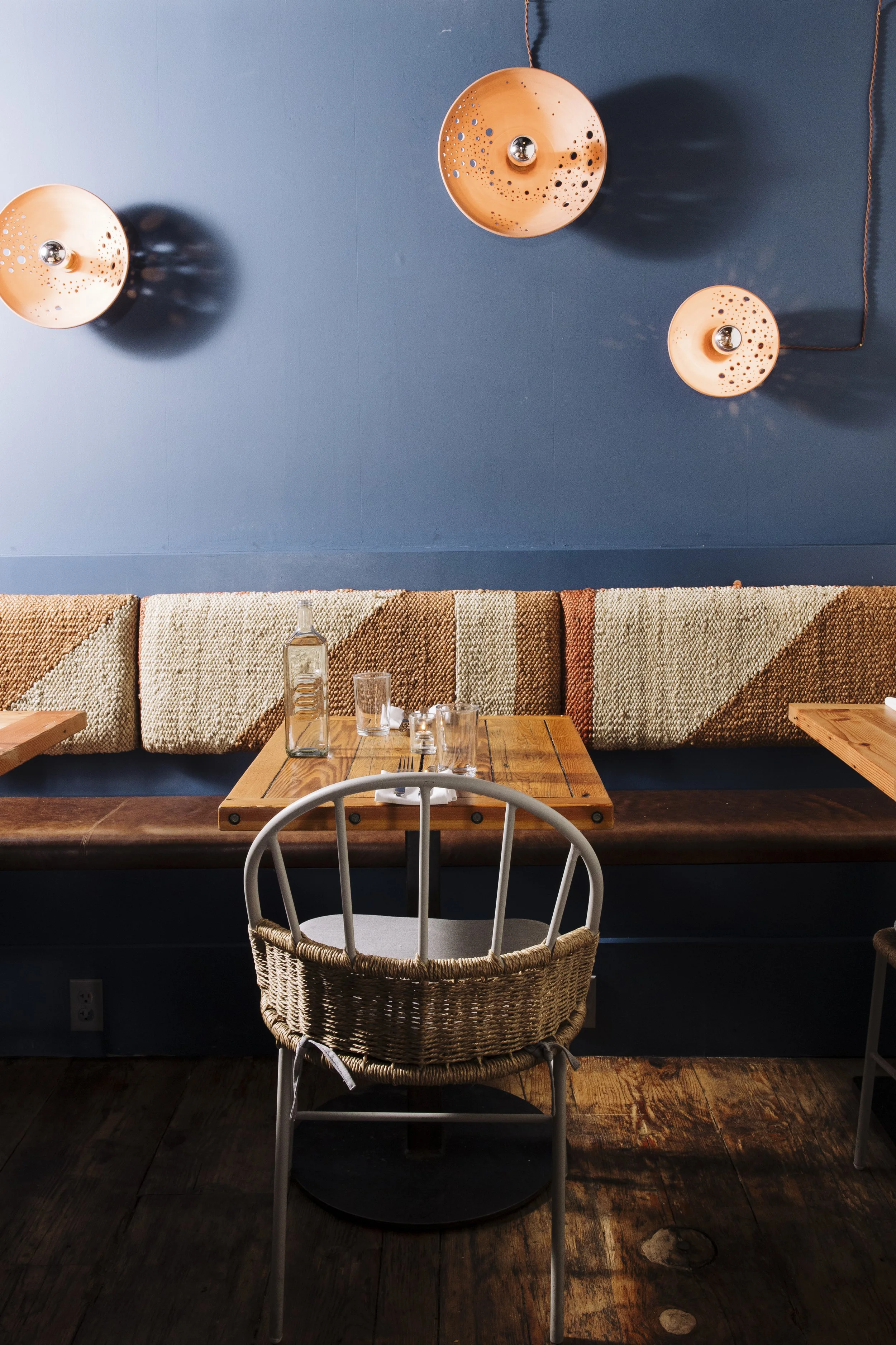 Interior view of a restaurant with a blue wall adorned with three hanging pendant lights, a wooden table set with a glass bottle and glasses, a woven-back chair in the foreground, and a long upholstered bench with a geometric pattern along the wall.