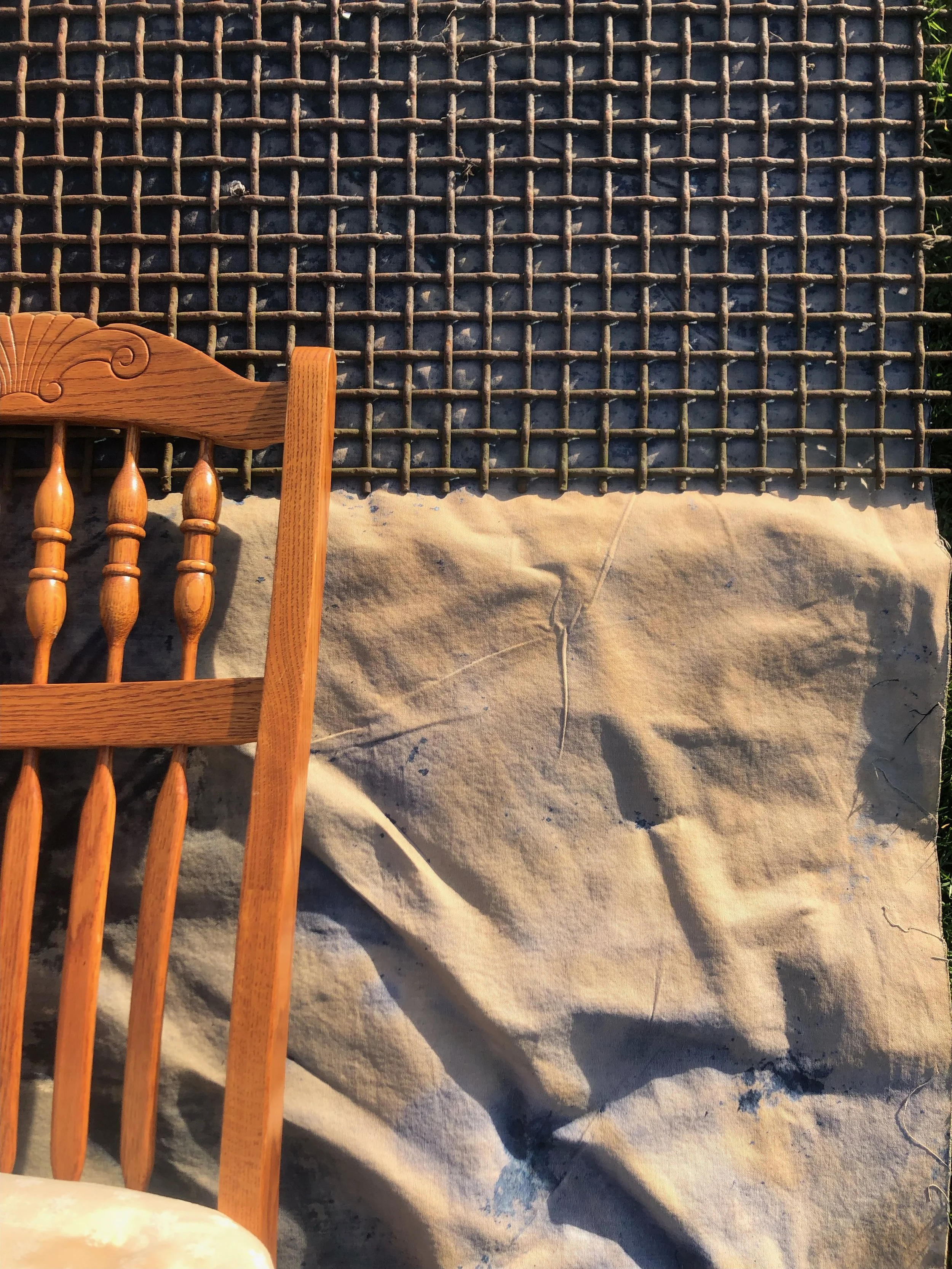 Part of a wooden chair with vertical spindles and carved decorative details, placed near a large fabric or paper surface with a grid pattern made of intersecting sticks.