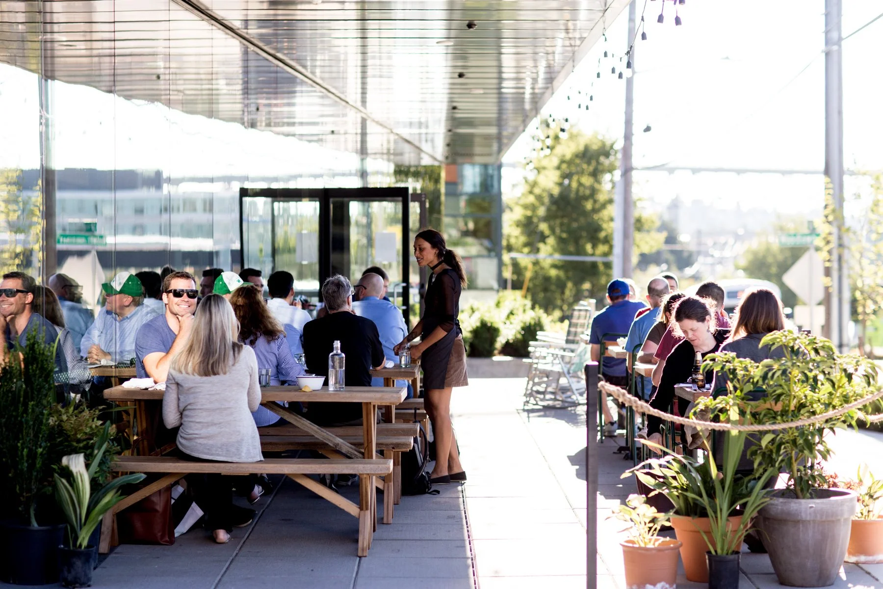 People dining outdoors at a restaurant with large potted plants and a reflective glass building in the background, under a covered patio with string lights.