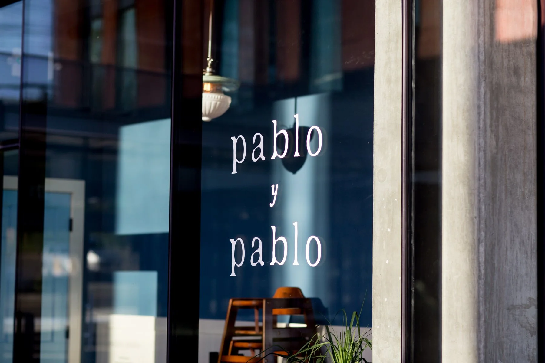 Glass window with the words 'pablo y pablo' written in white text, reflecting interior furniture and a plant outside.