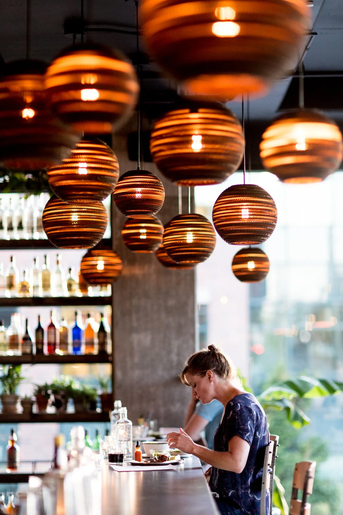 Woman sitting at a bar counter with hanging round orange and brown striped pendant lights above, and shelves of bottles behind her, enjoying a meal.