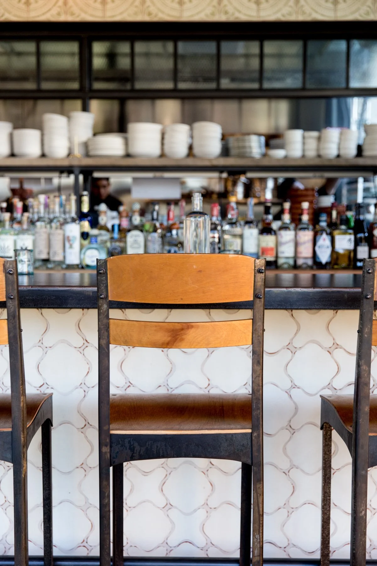 View of a bar or restaurant interior with chairs and a bar counter with bottles in the background. Shelves with white bowls are on the upper part of the image.