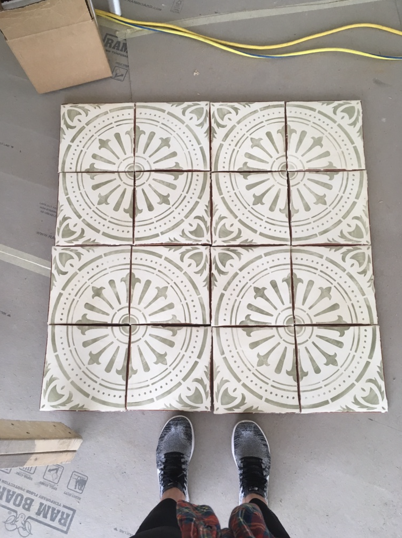 A person wearing black sneakers and plaid shorts standing on a gray concrete floor in front of a stack of decorative ceramic tiles with a white and beige circular pattern.