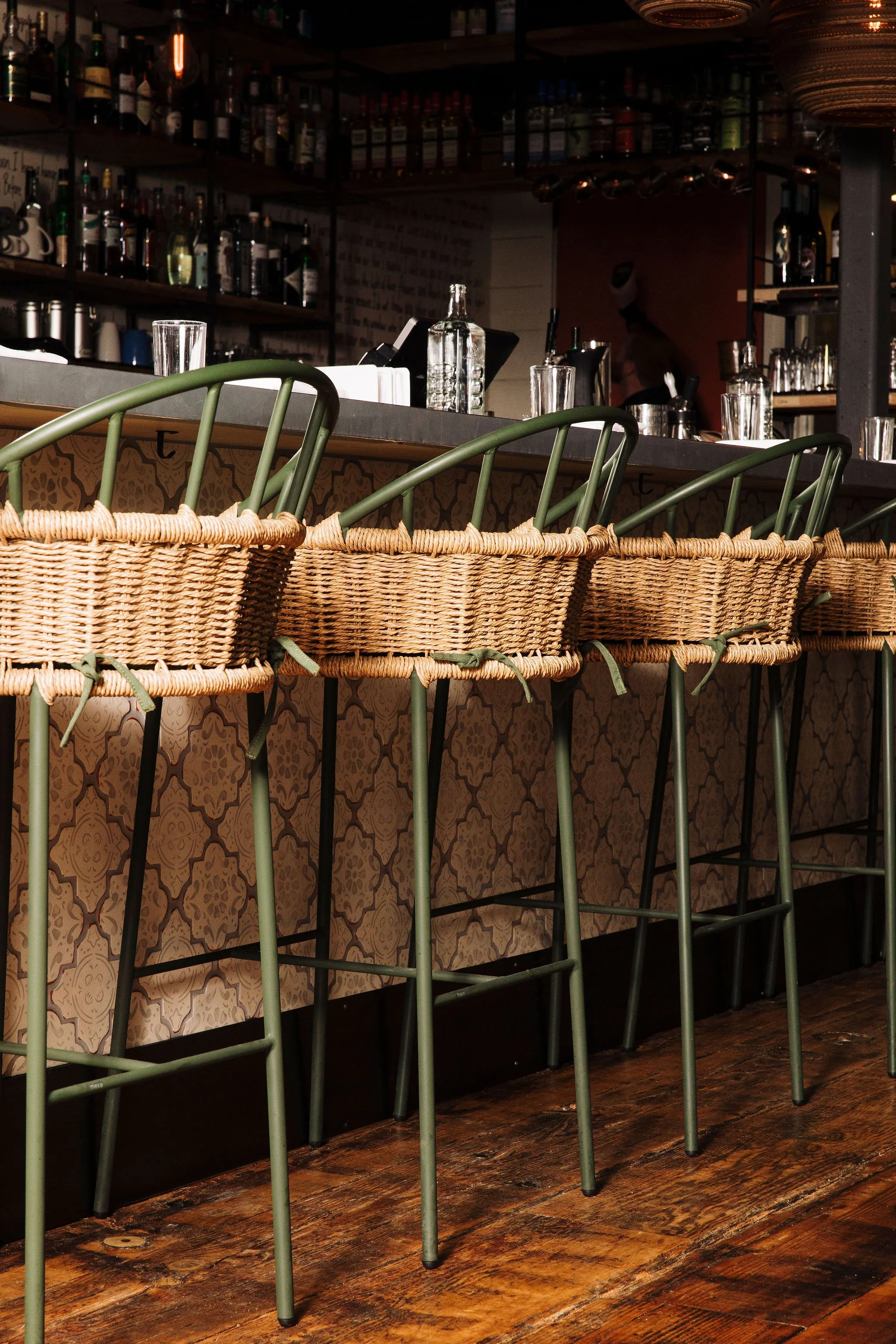 A bar with a dark counter, glassware, bottles on shelves, and four green metal bar stools with woven seats in the foreground, on a dark wooden floor.
