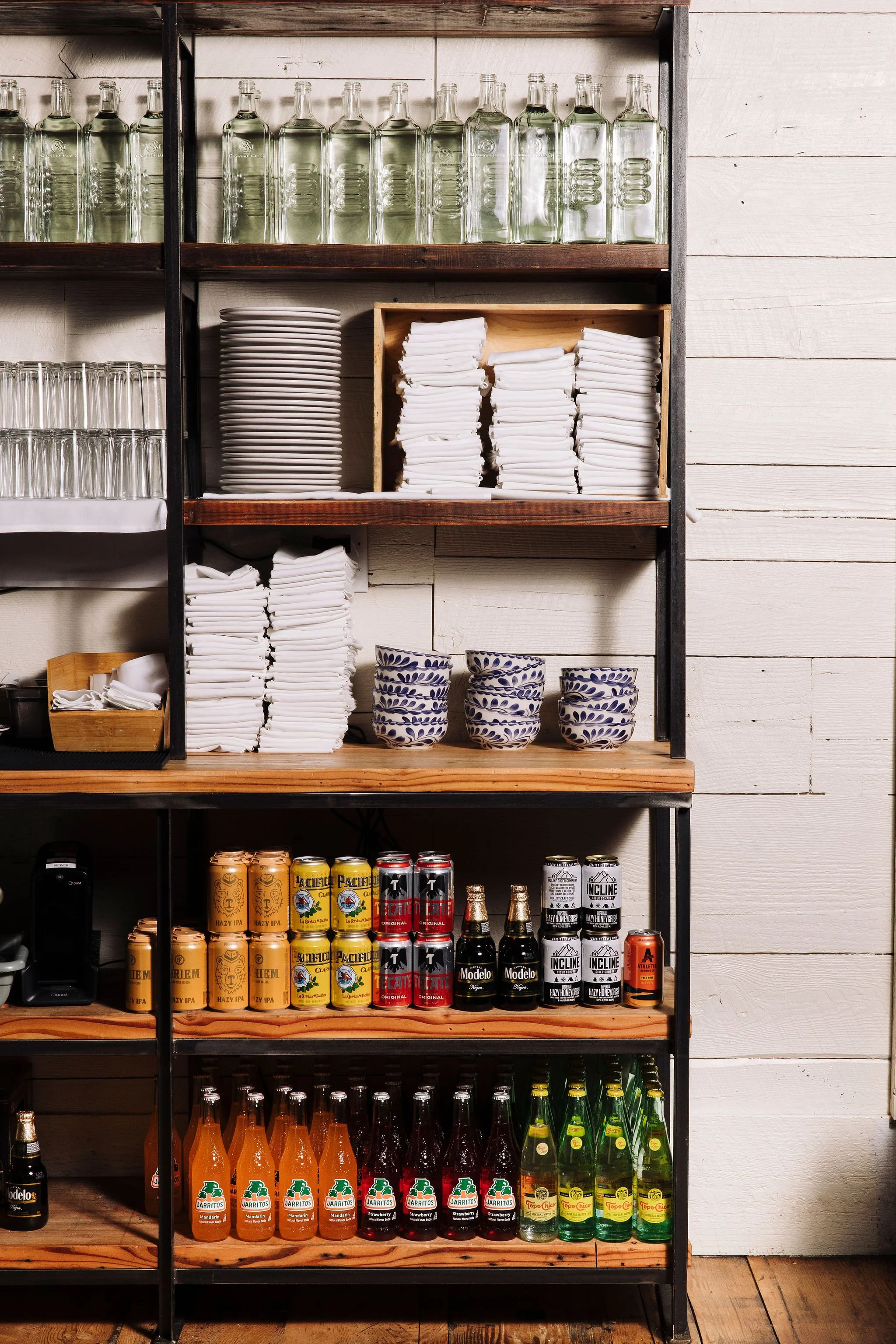 Shelving unit with glass bottles, plates, and canned beverages, including beer and juice, arranged on wooden shelves against a white wall.