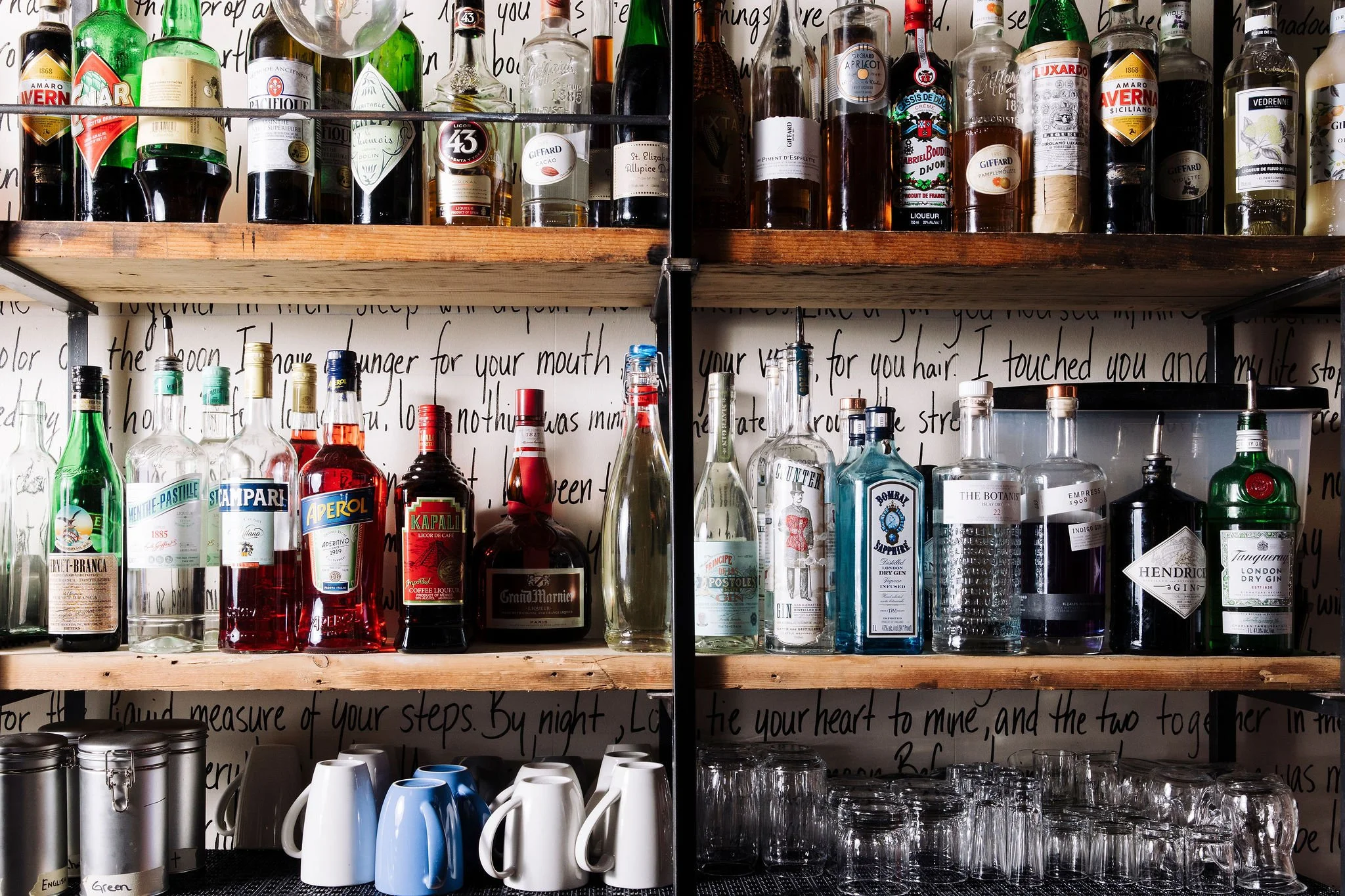Shelves filled with various bottles of alcohol including gin, vodka, whiskey, and liqueurs, along with mugs and glasses underneath, in a bar setting.