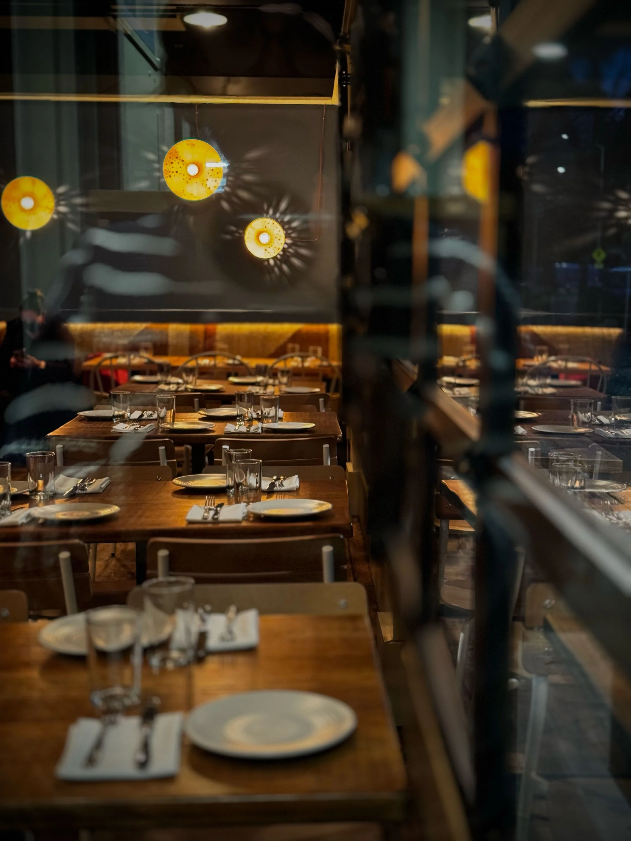 Empty restaurant dining area with tables set with plates, glasses, and silverware, seen through a window with reflections of three hanging yellow pendant lights.