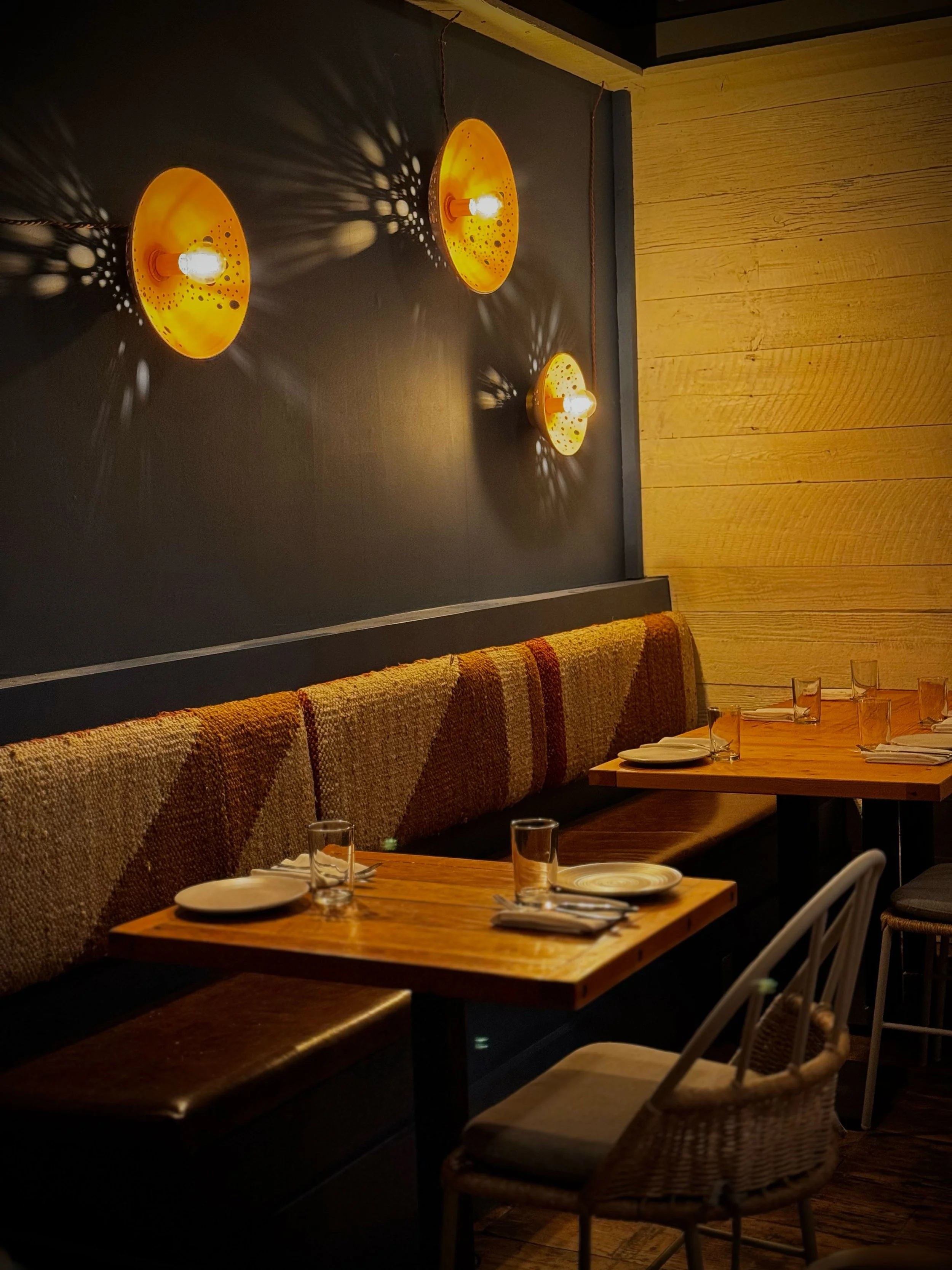 A cozy restaurant corner with wooden tables set with plates, glasses, and napkins, a cushioned bench with a textured, patterned fabric, chairs, and a dark wall with yellow-orange lamps casting patterned shadows.