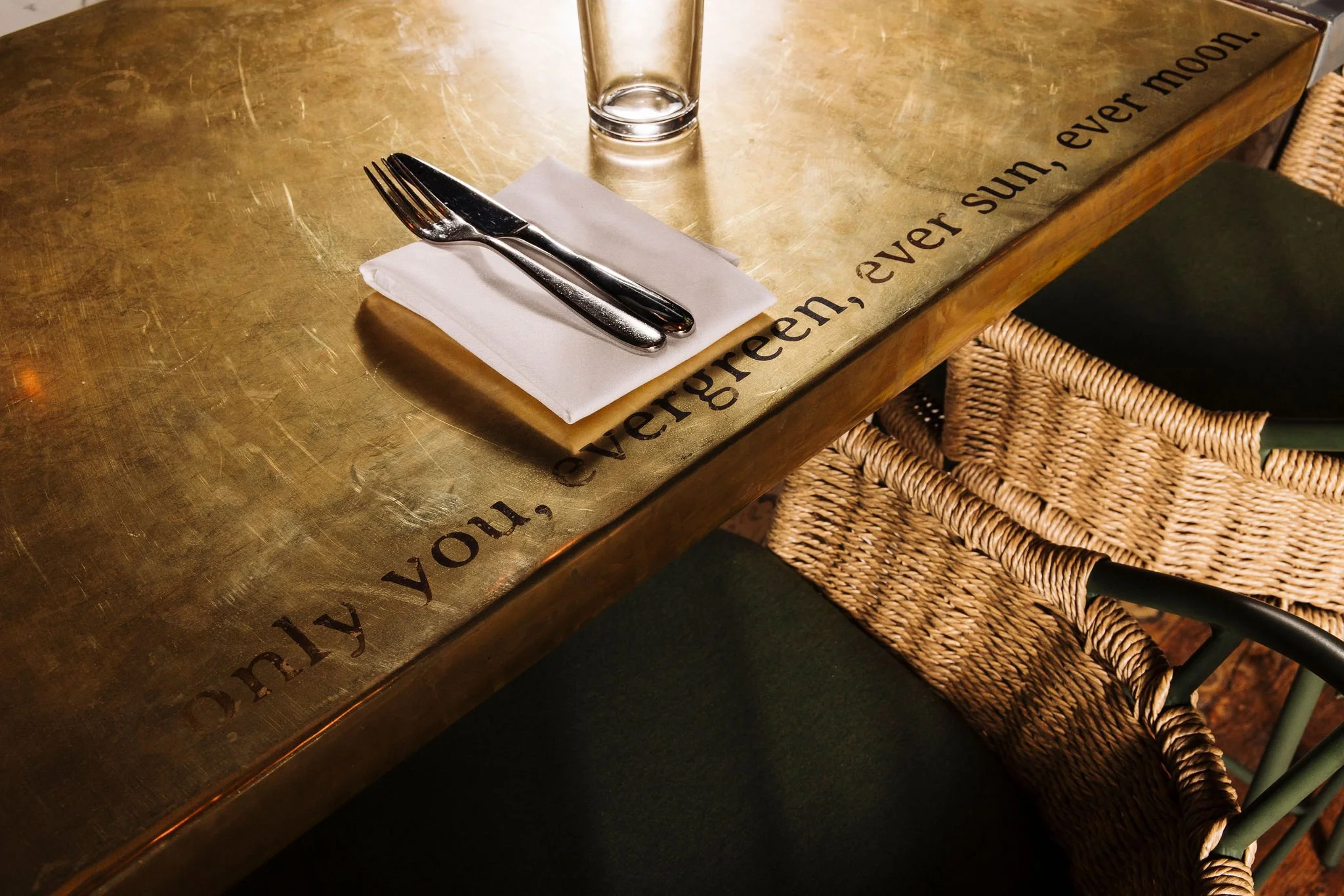 A table setting with a fork and knife placed on a white napkin, a clear glass, and a metallic placemat with the quote 'The greener sun, even moon' written on the edge, all on a wooden table with wicker chairs nearby.