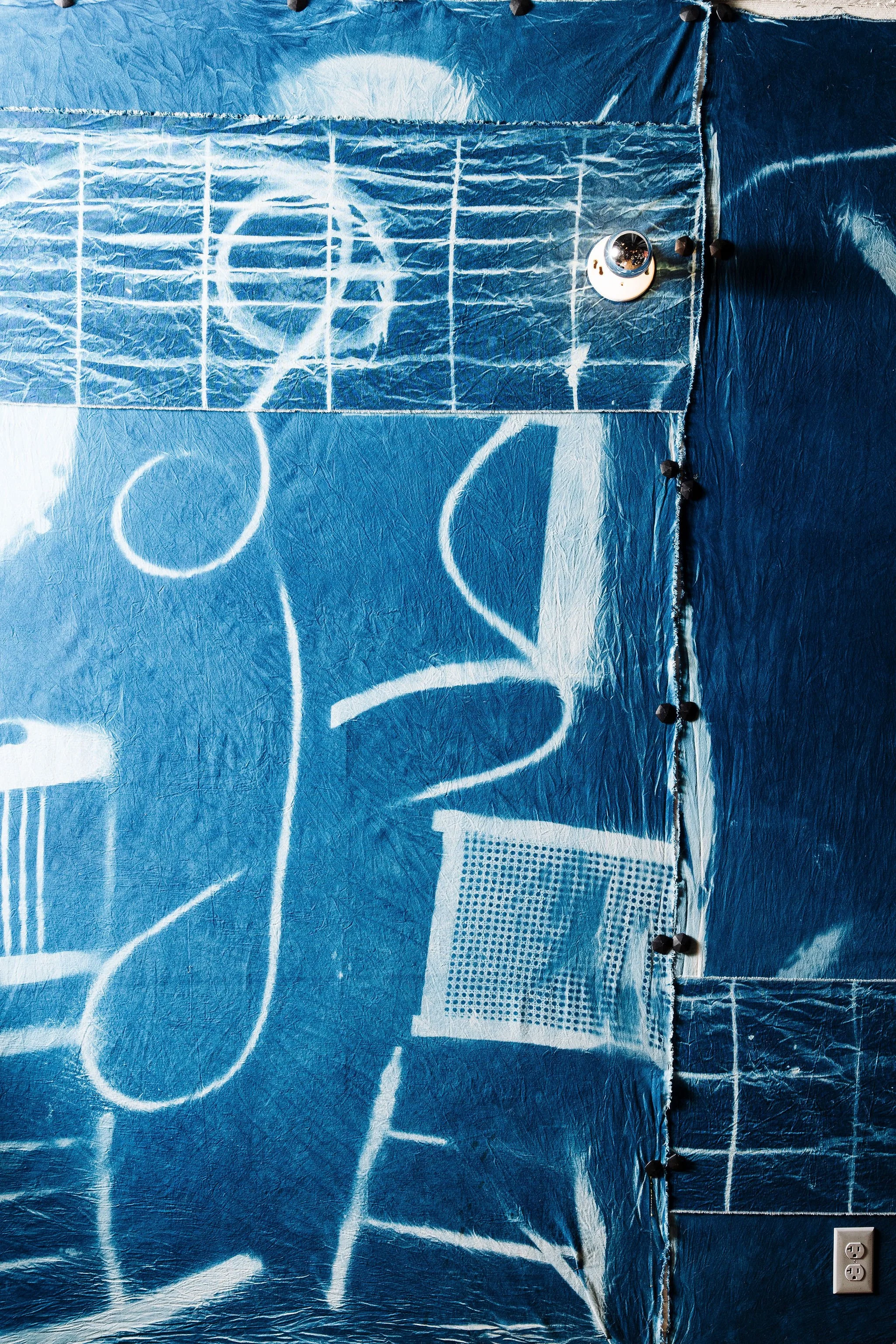 Close-up of a blue wall with a mural or painted design featuring white chalk-like marks outlining a basketball hoop, backboard, and some abstract shapes. There are black push pins along the right edge and a white electrical outlet at the bottom right