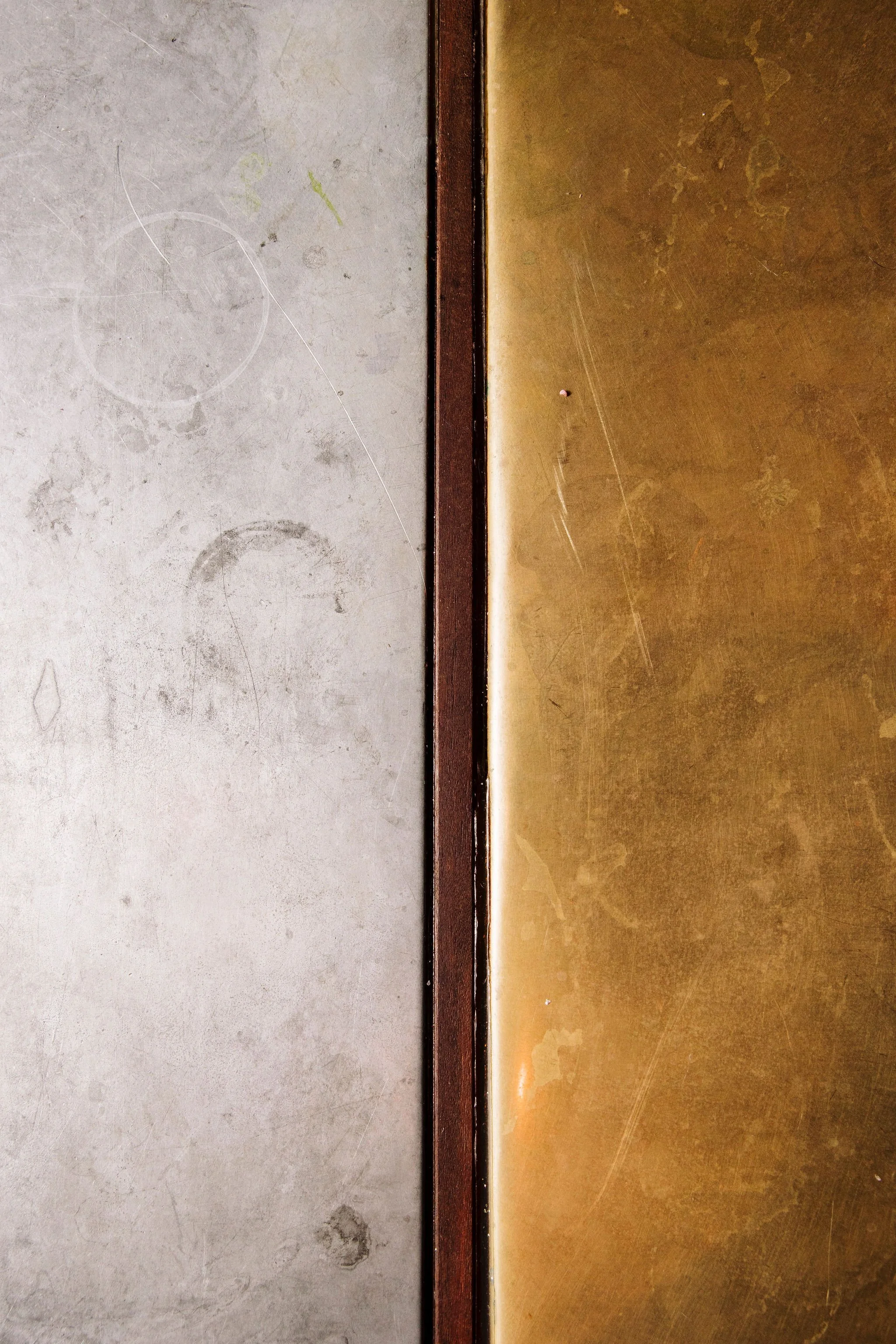 A close-up of two side-by-side surfaces separated by a thin trim. The left surface is gray with scuff marks, circular stains, and scratches. The right surface is golden with a textured, slightly worn appearance. The trim between the surfaces is dark 