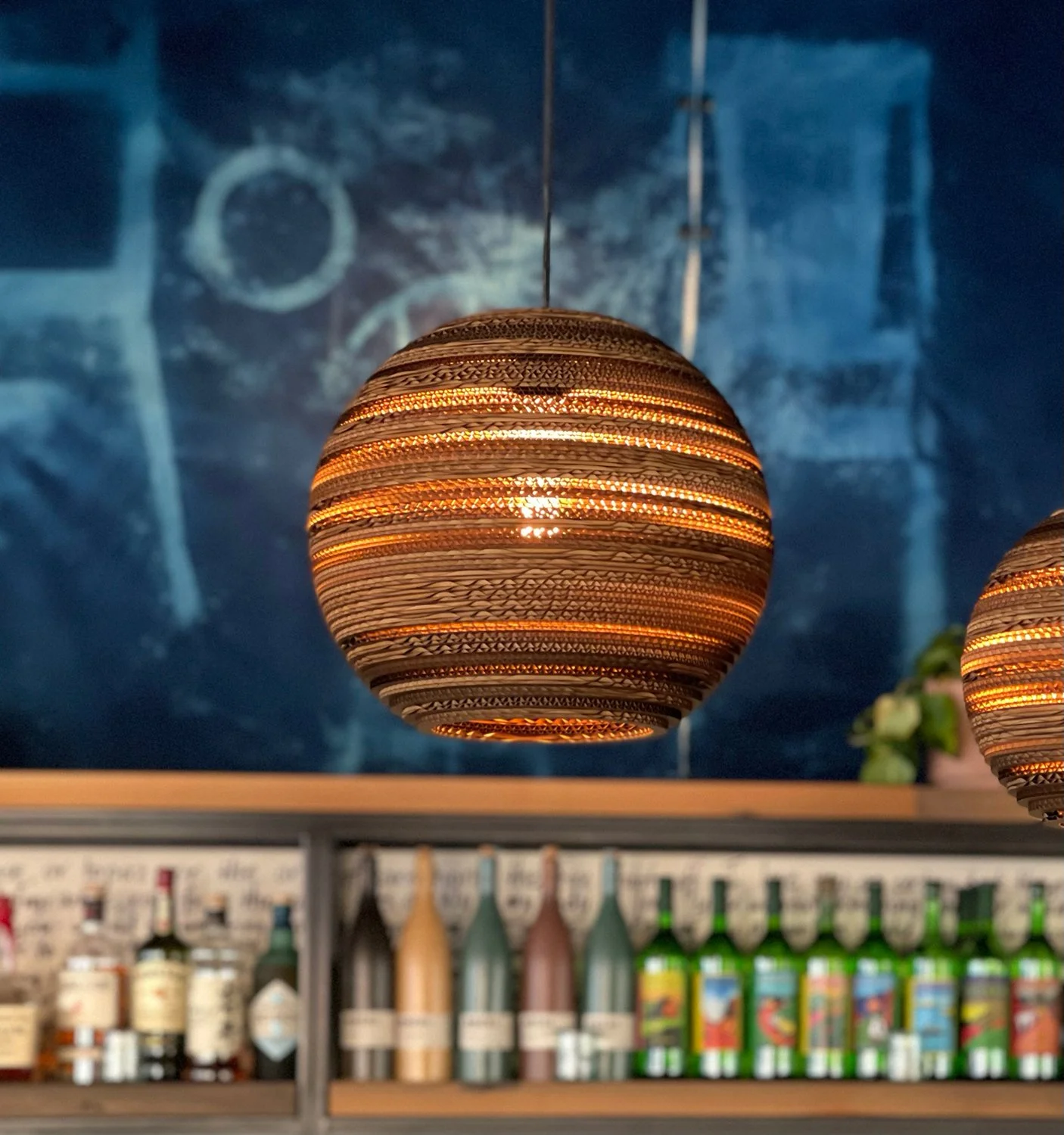 A woven, round, hanging pendant light glowing with warm light in front of a blue wall with abstract circle and line art
