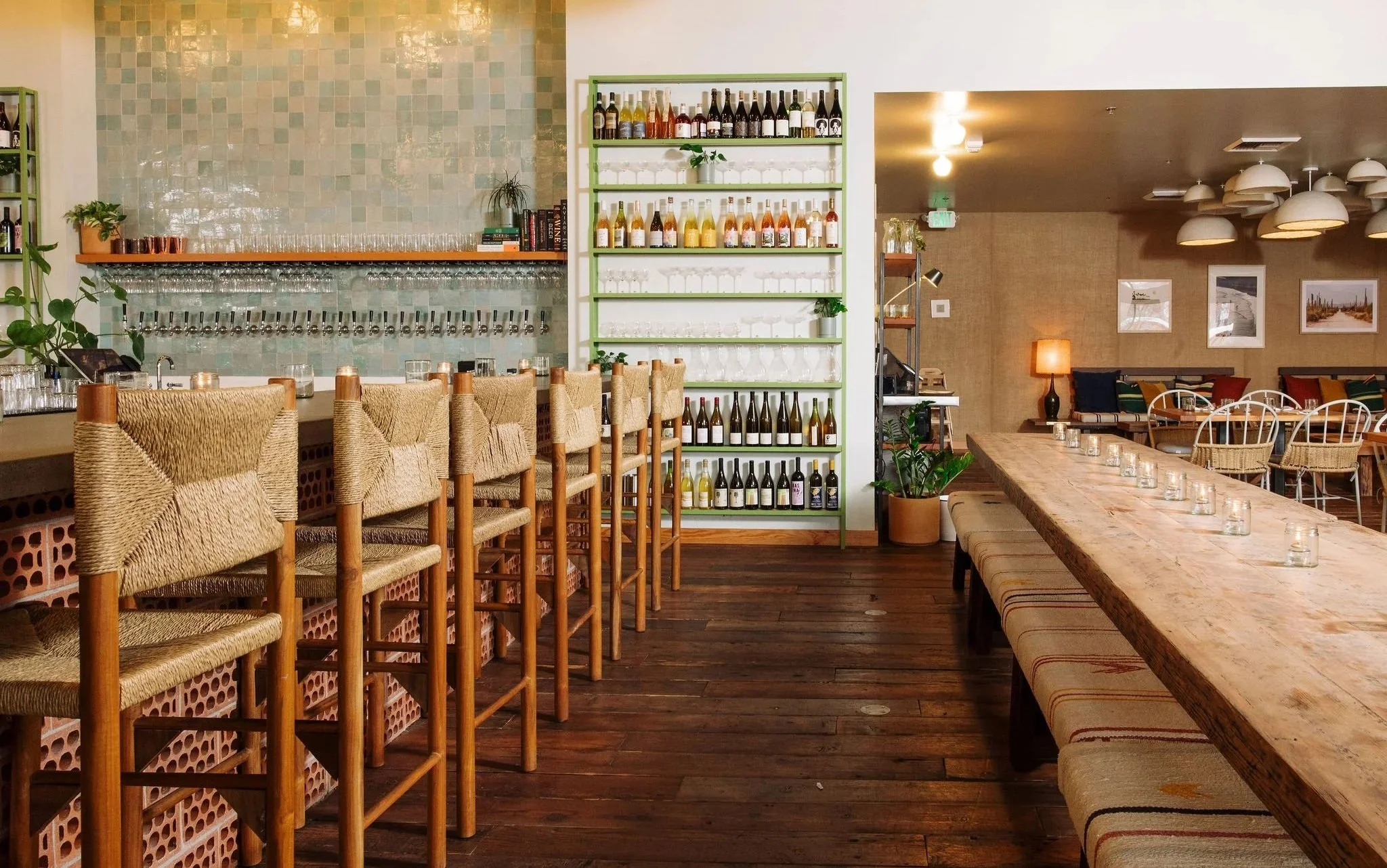 Interior of a restaurant or bar with a long wooden table, woven chairs, a shelving unit with wine bottles, and a bar area with glasses and plants.