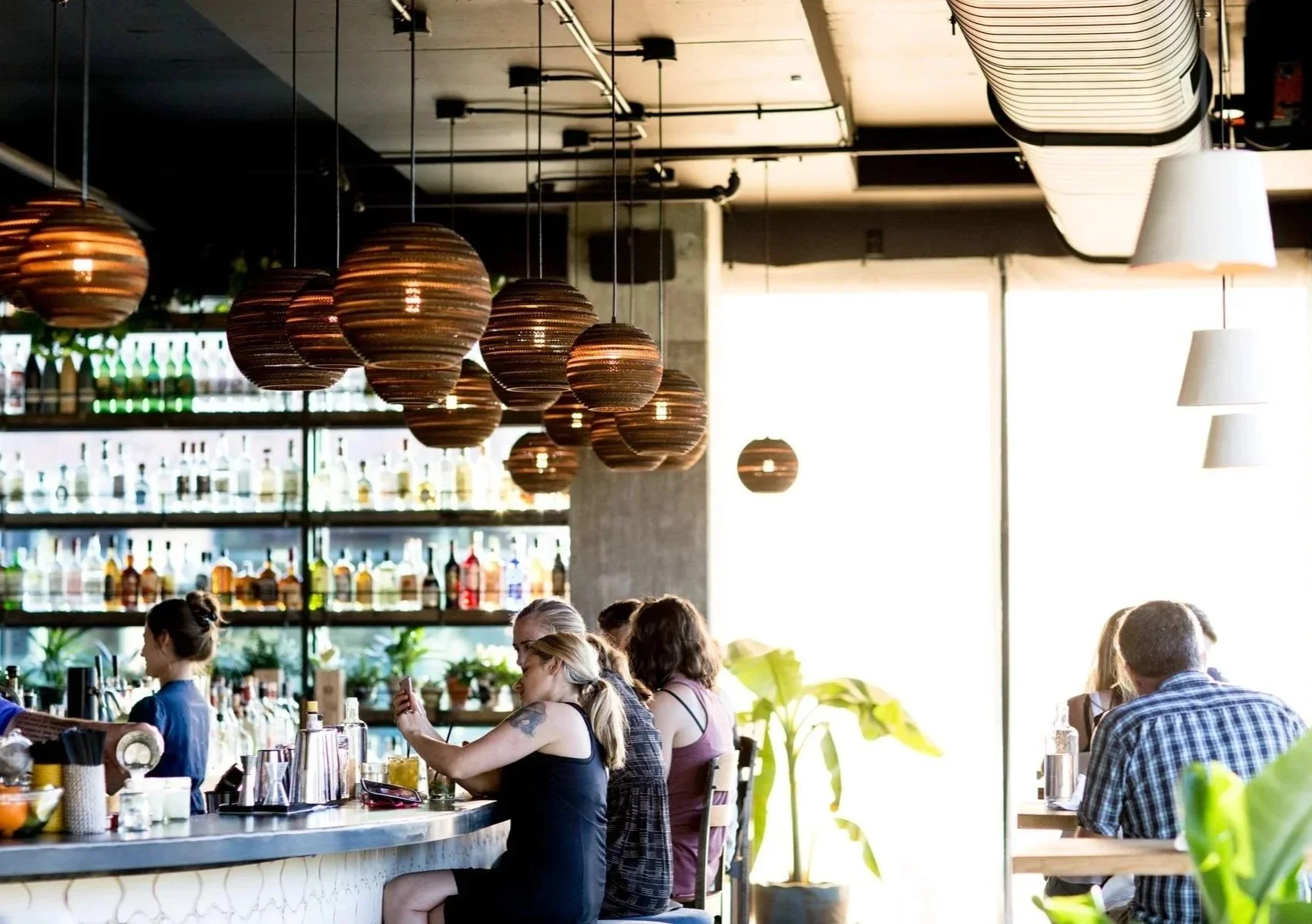 People sitting at a bar in a modern restaurant with hanging wooden sphere light fixtures and shelves of liquor bottles in the background.
