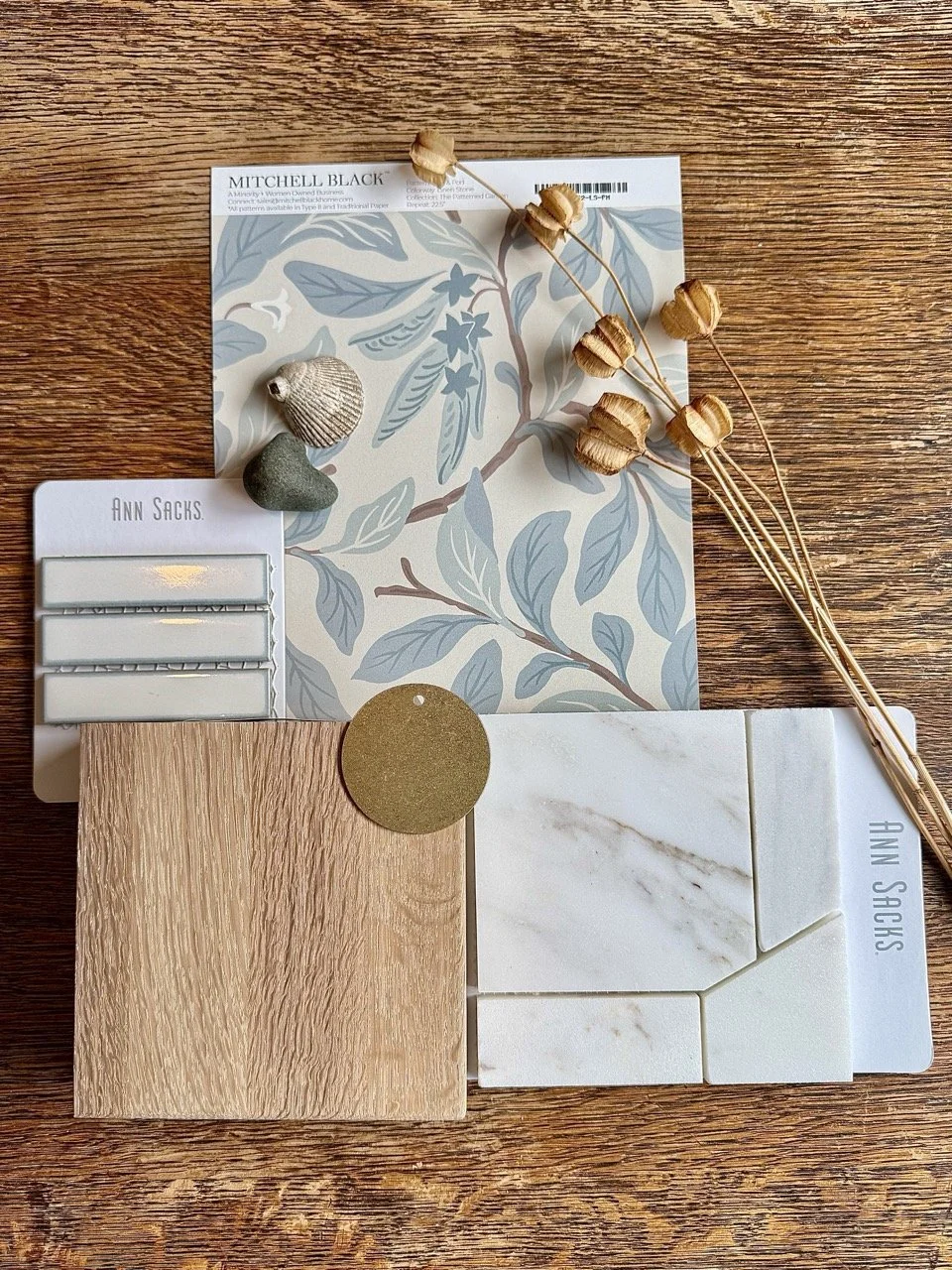 Decorative display with dried flowers, shell and stone, patterned wallpaper, sample tiles, and a sample wood panel on a wooden surface.