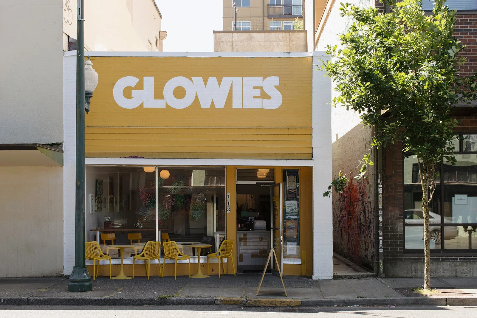 Front view of a storefront with a large yellow sign reading 'GLOWIES' in white letters. The store has a glass window with yellow chairs and tables outside. There is a tree on the right side of the storefront and a streetlamp on the left.