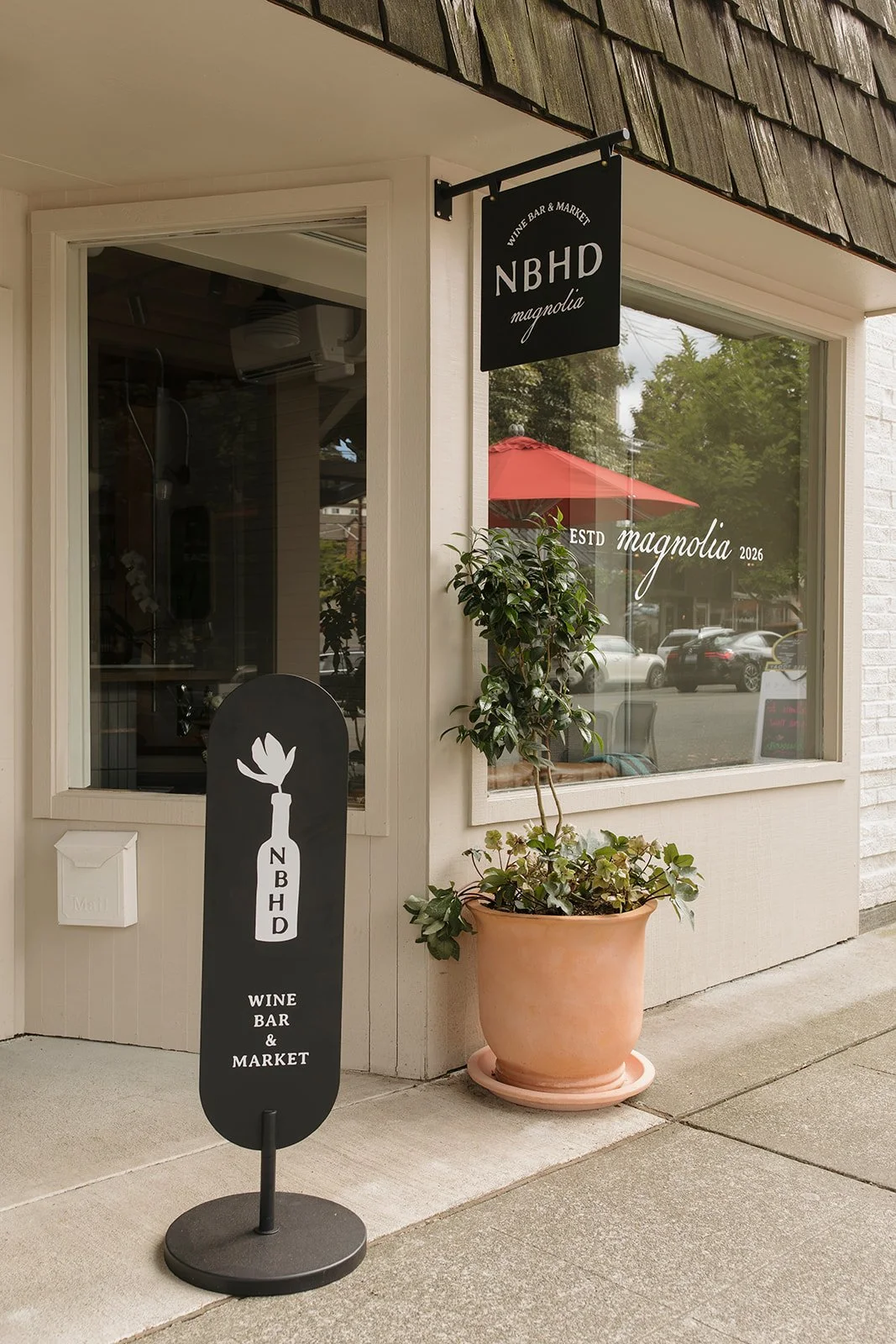 The exterior of a shop named NBHD Magnolia, a wine bar and market, with a black sign hanging outside. There is a large potted plant next to the window and a black sandwich board sign on the sidewalk. The shop window has the name 'Magnolia' and 'ESTD 