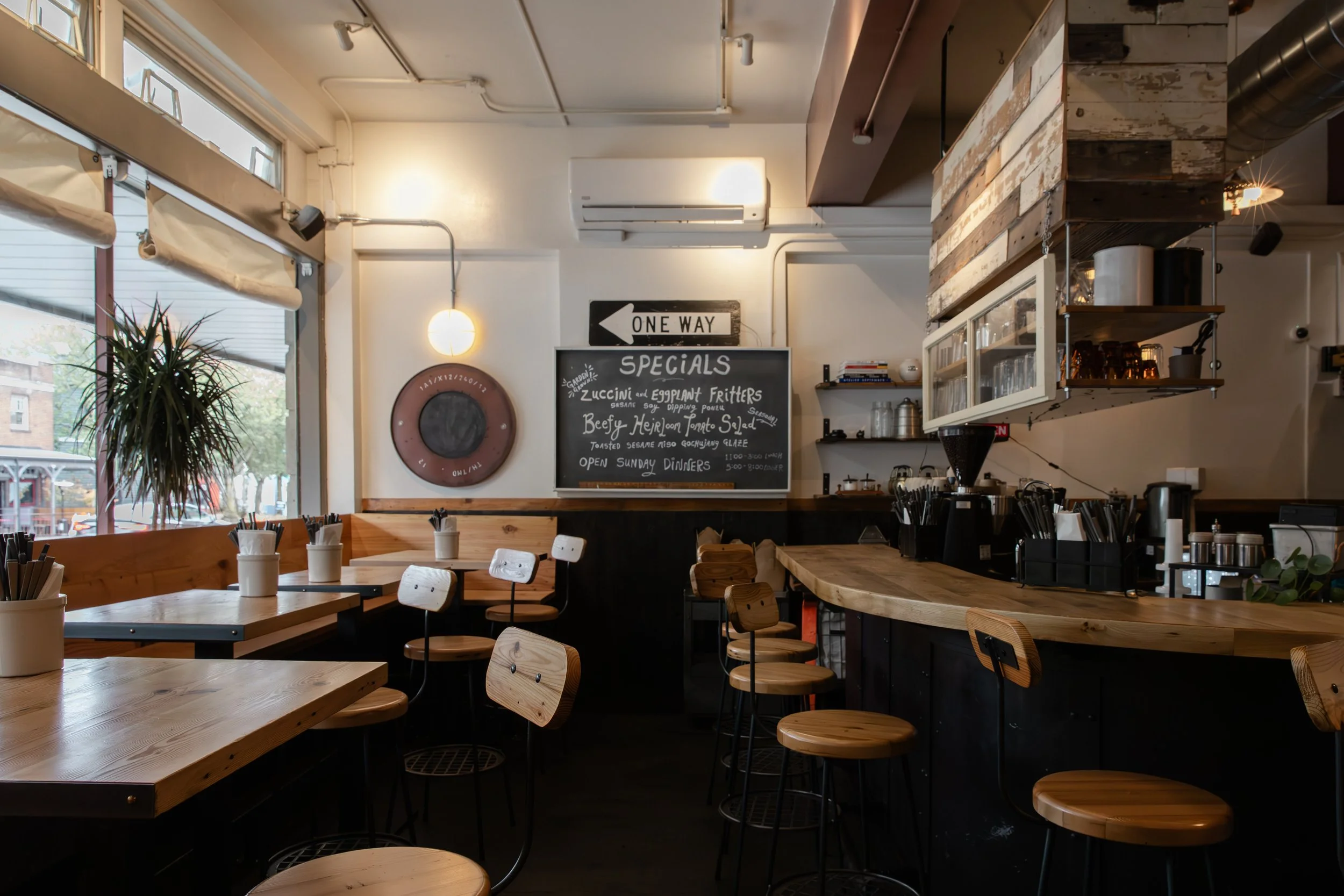 Interior of a casual cafe with wooden tables, chairs, a chalkboard menu listing specials, and large windows letting in natural light.