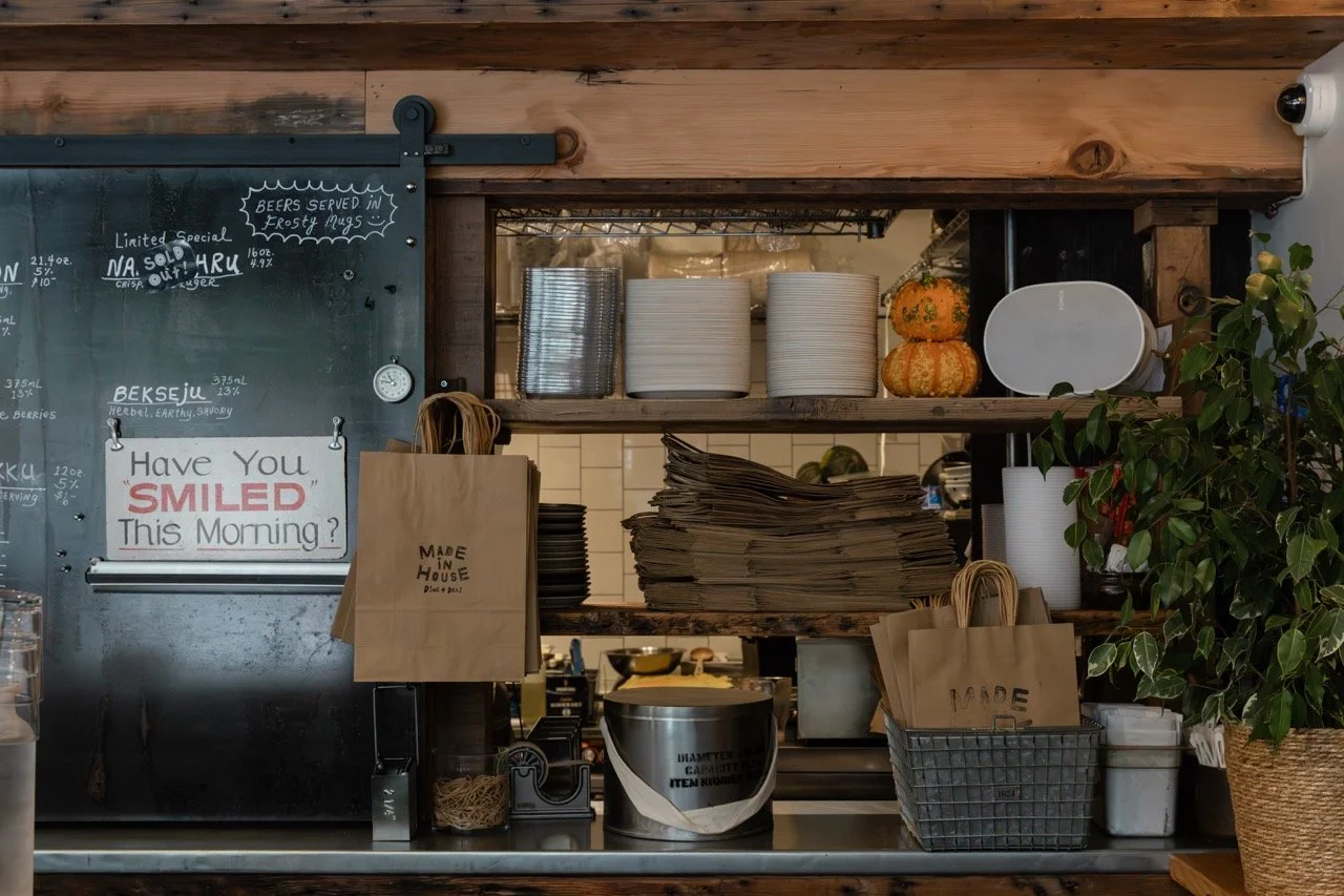 Kitchen shelf with stacked white plates, paper napkins, paper bags, and a potted plant, with a chalkboard menu in the background.