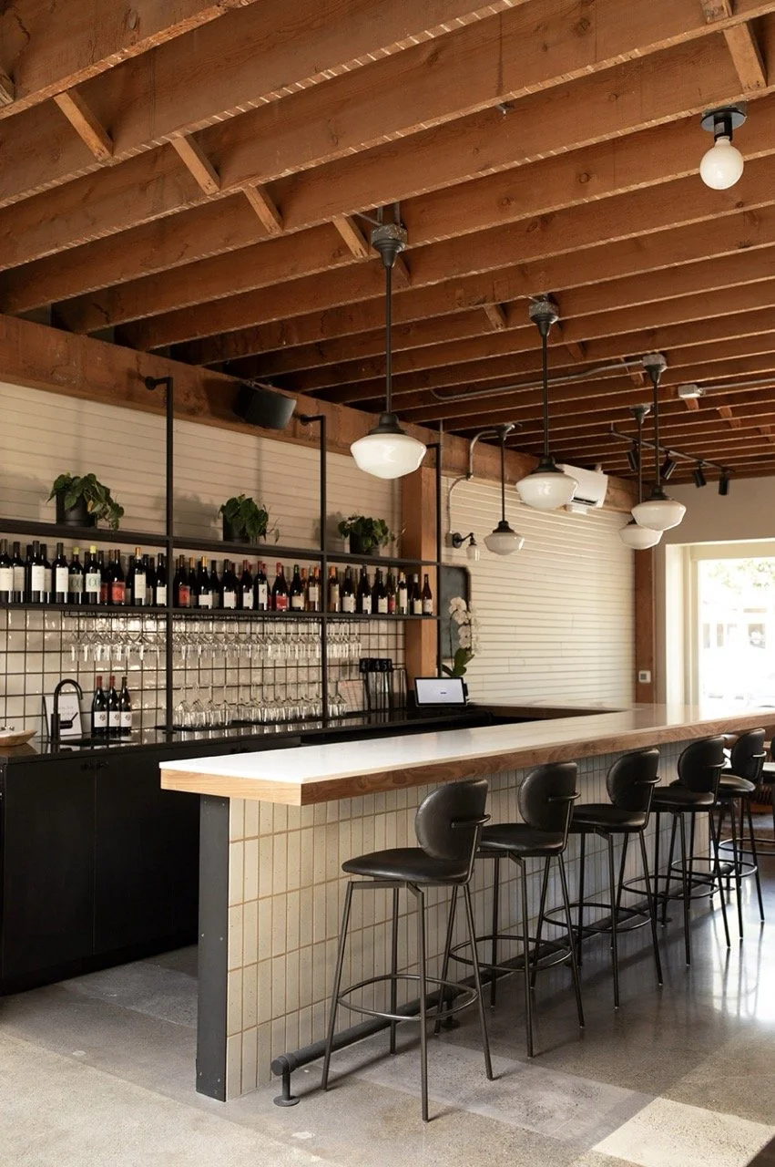Interior of a modern bar with a long white countertop, black barstools, exposed wooden ceiling beams, hanging pendant lights, and a shelf with wine bottles and glasses behind the bar.