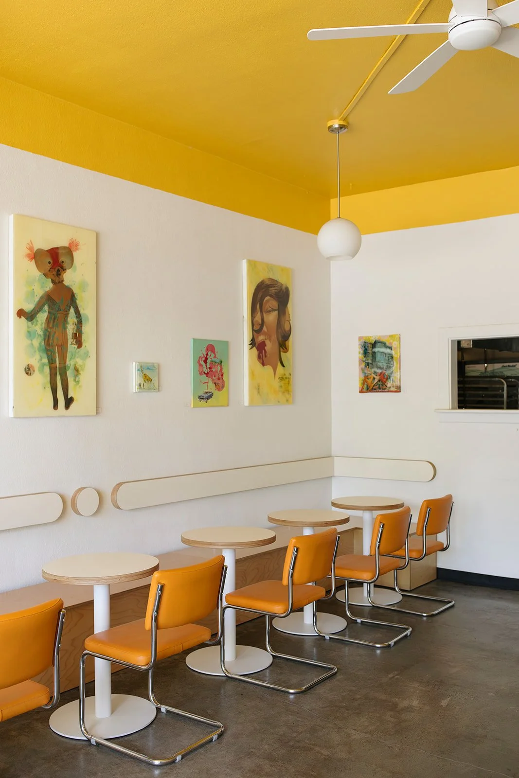 Empty restaurant or cafe interior with yellow ceiling, white walls, and orange chairs at light wooden tables. Artworks hang on the wall, and there is a ceiling fan and a hanging light.