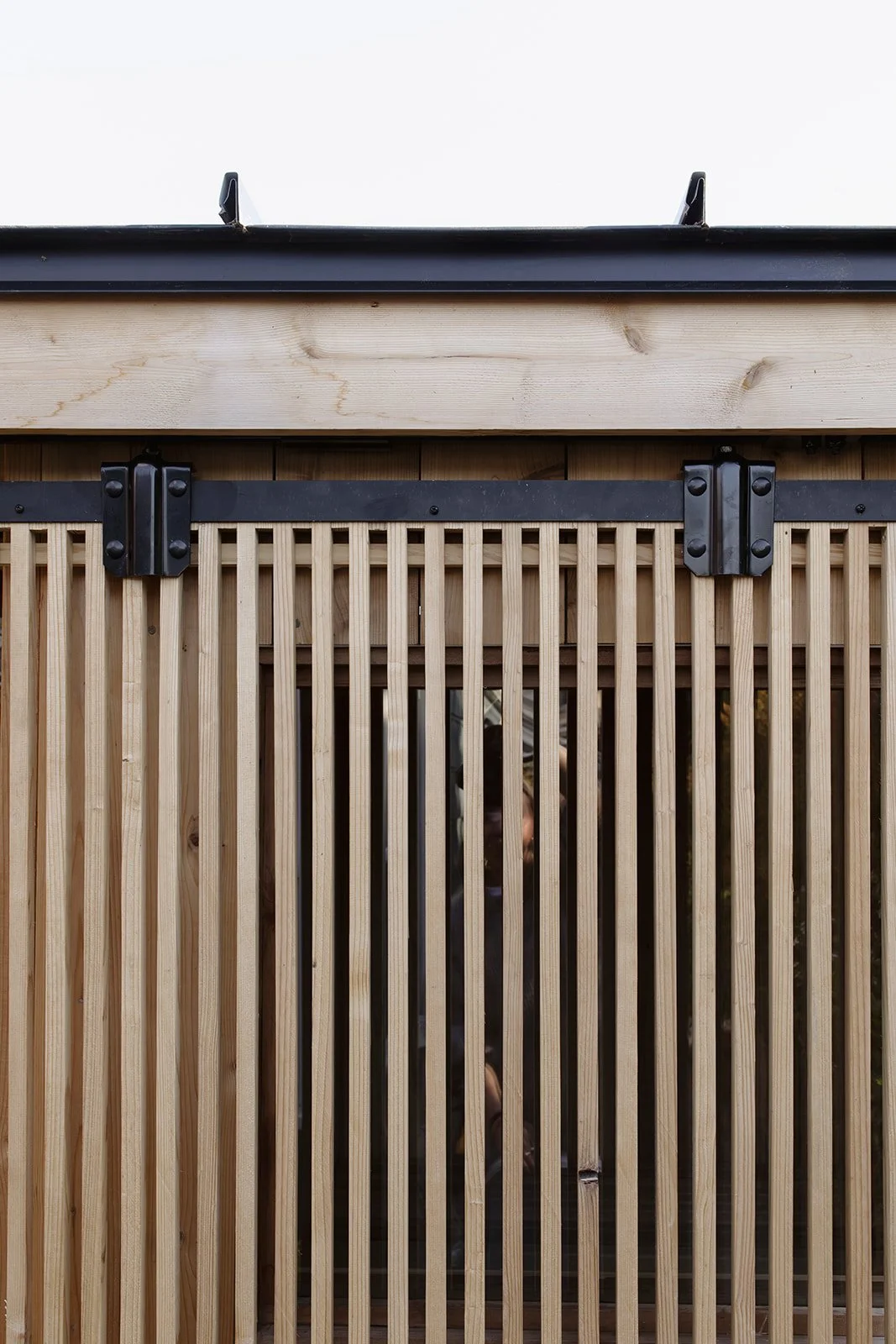 A wooden structure with vertical slats and black hardware hinges, seen through a glass panel with a reflection of a person taking the photo.