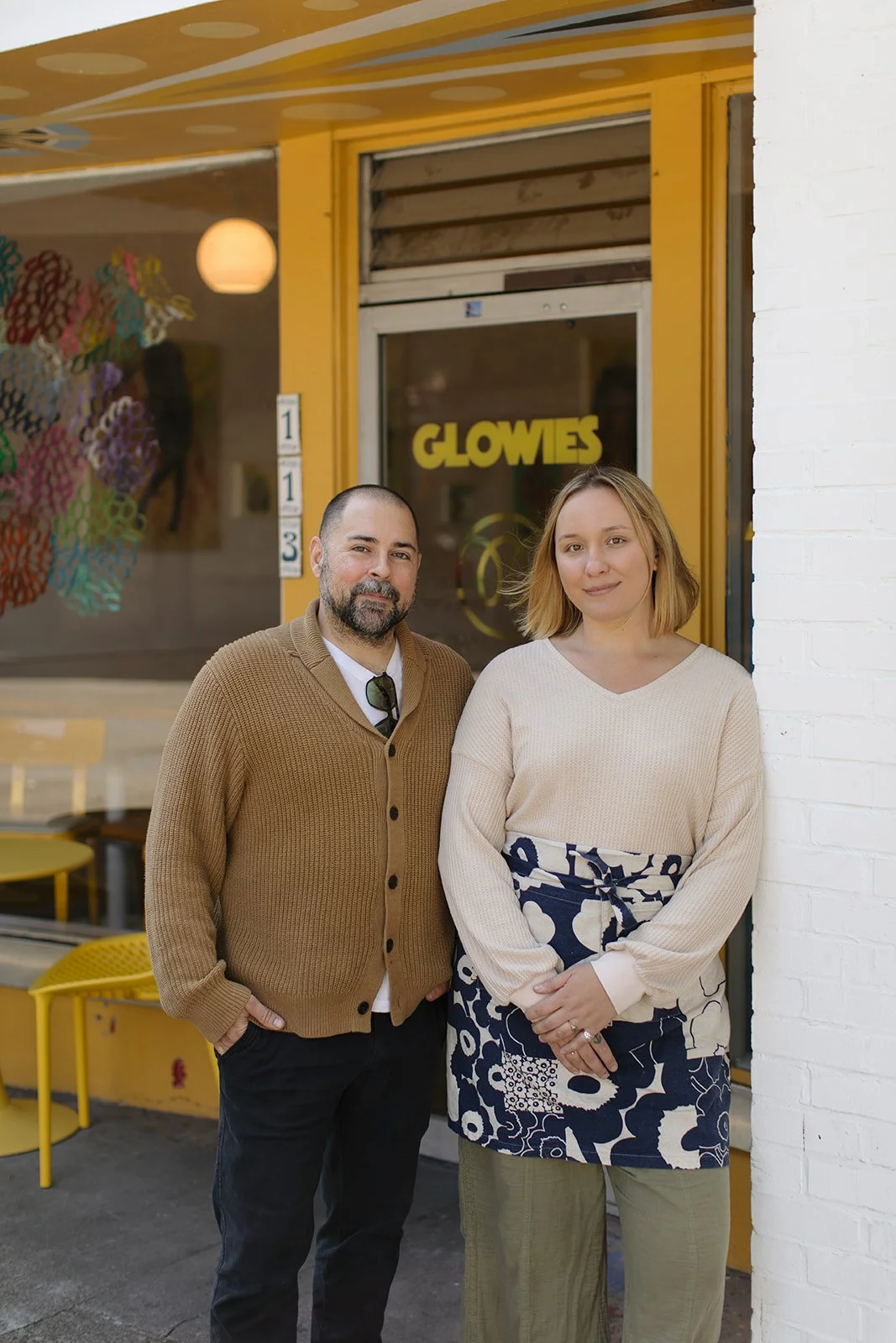 A man and a woman standing outside a store with a yellow facade. The store is named 'GLOWIES' as seen from the yellow sign on the window. The man has a beard, wears a brown cardigan, white shirt, and sunglasses hanging from his shirt collar. The woma