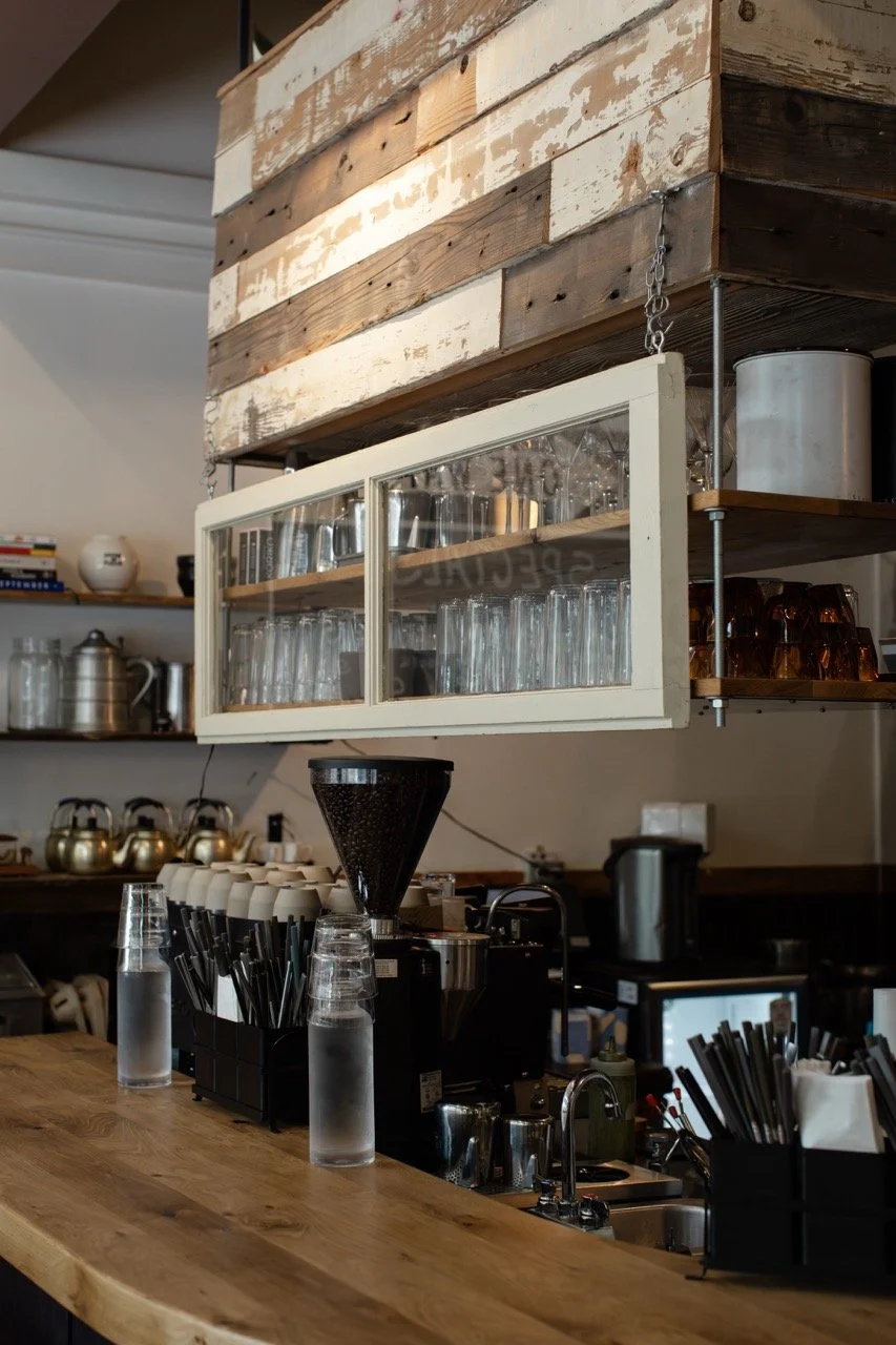 Inside a coffee shop, showing a wooden counter with water glasses, a coffee grinder, and utensils, and shelves with glasses and cups above.