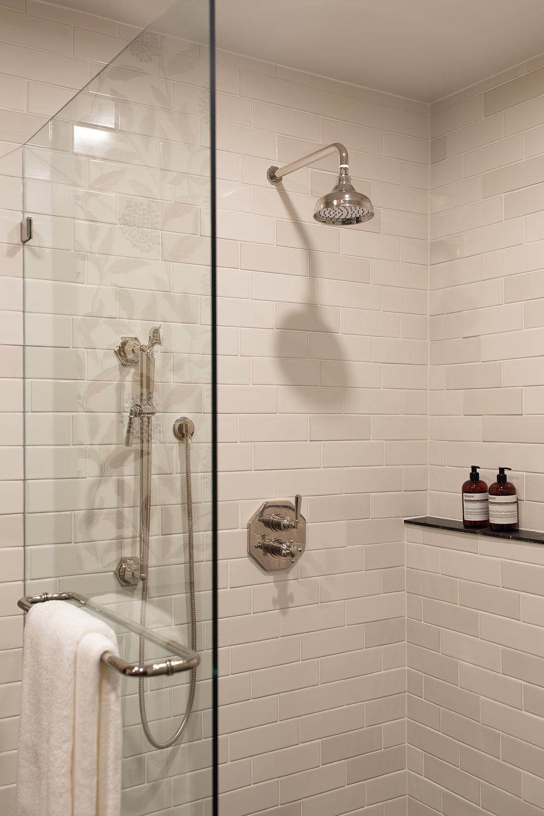 A modern shower with a handheld showerhead, a fixed overhead shower, white subway tiles, a towel rack with a white towel, and two brown bottles on a black shelf.