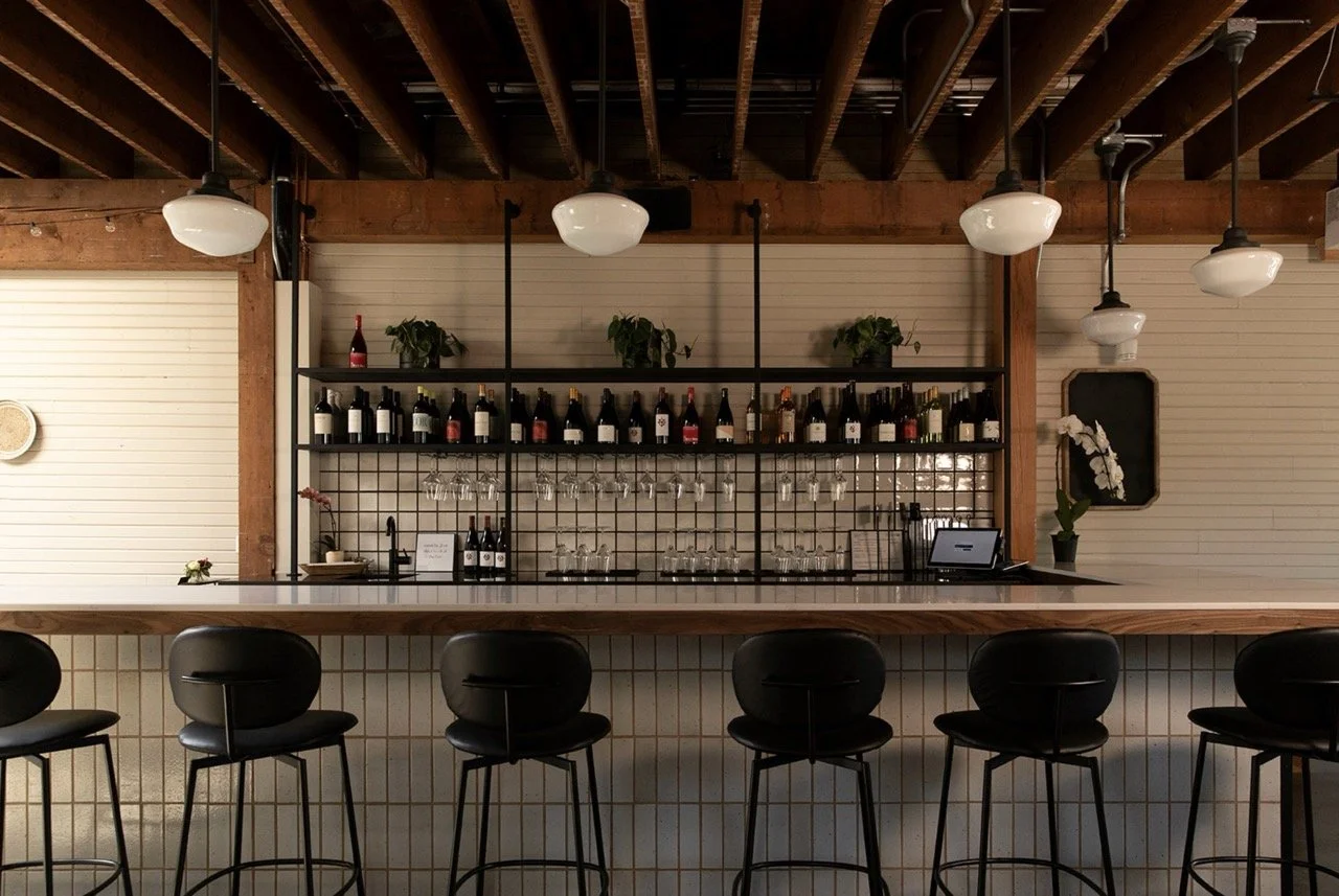 Interior of a wine bar with a wooden bar counter, black chairs, wine bottles and glasses on a shelf behind the bar, and hanging pendant lights.