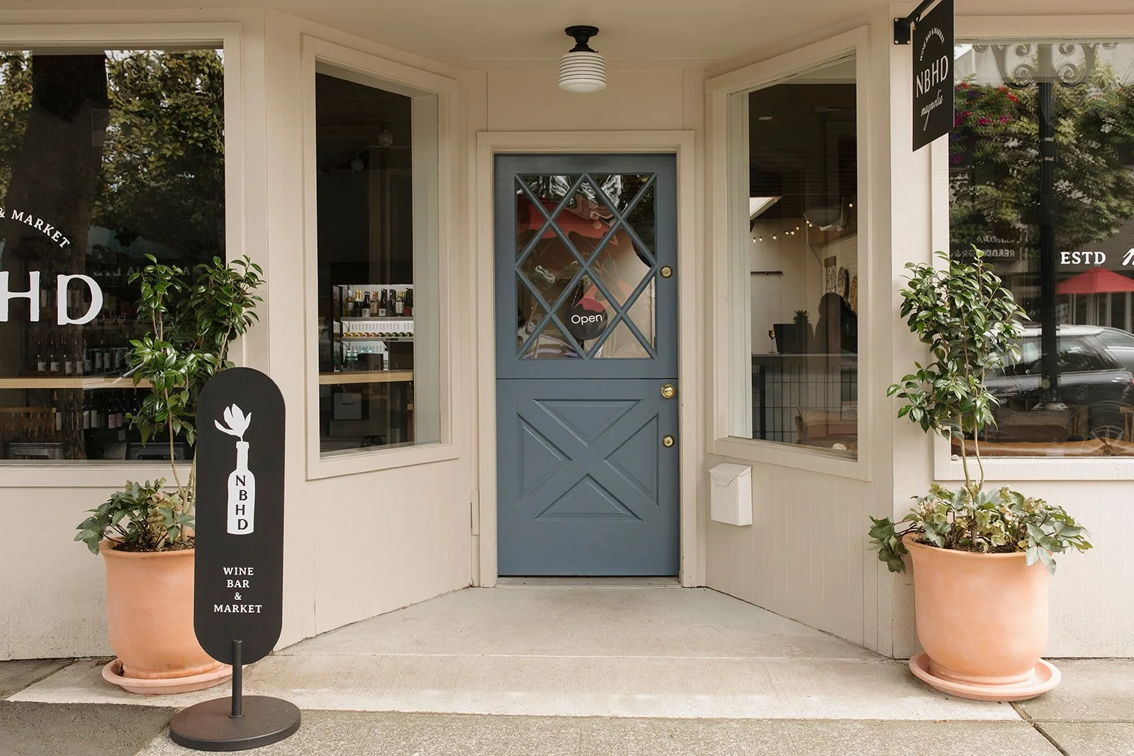 Entrance to a wine bar and market with a blue door, two large potted plants flanking the doorway, and a black signboard on the sidewalk displaying the establishment's name and offerings.
