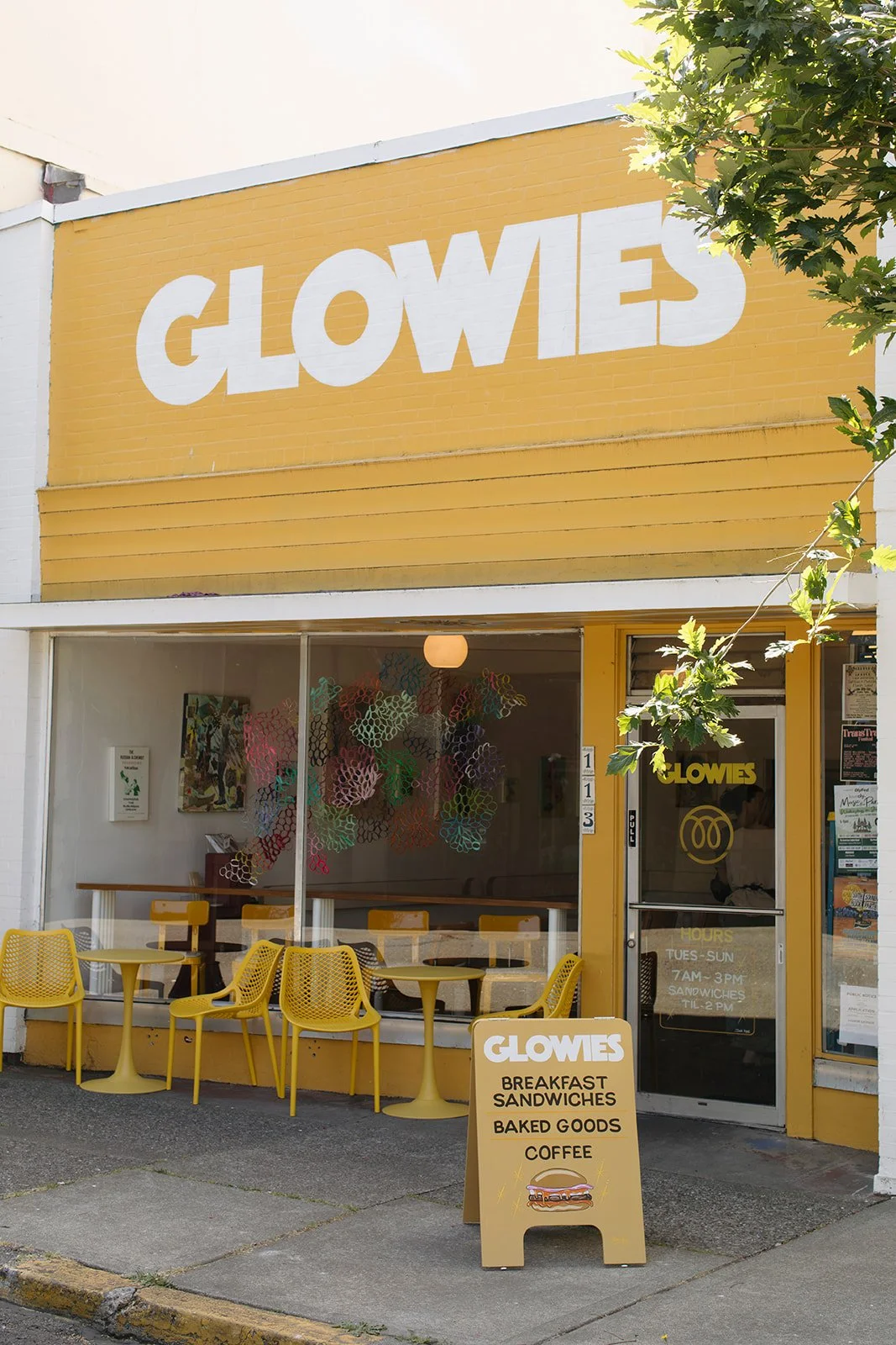 Yellow storefront with large white sign that reads 'GLOWES'. Inside, there are yellow chairs and tables, decorated with colorful paper circles. Outside, a sandwich board advertises breakfast sandwiches, baked goods, and coffee.