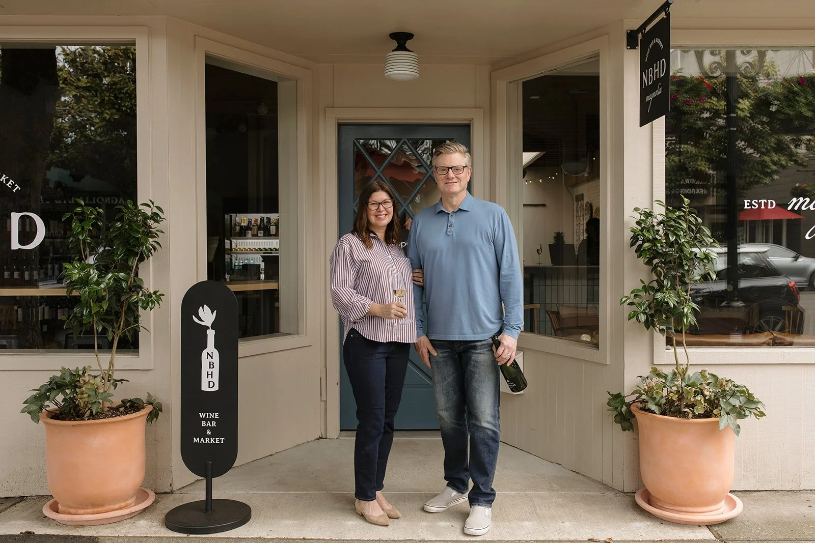 Two people standing outside a wine bar and market, holding wine glasses and a bottle, smiling at the camera, with potted plants on either side and a sign that reads 'Wine Bar & Market'.