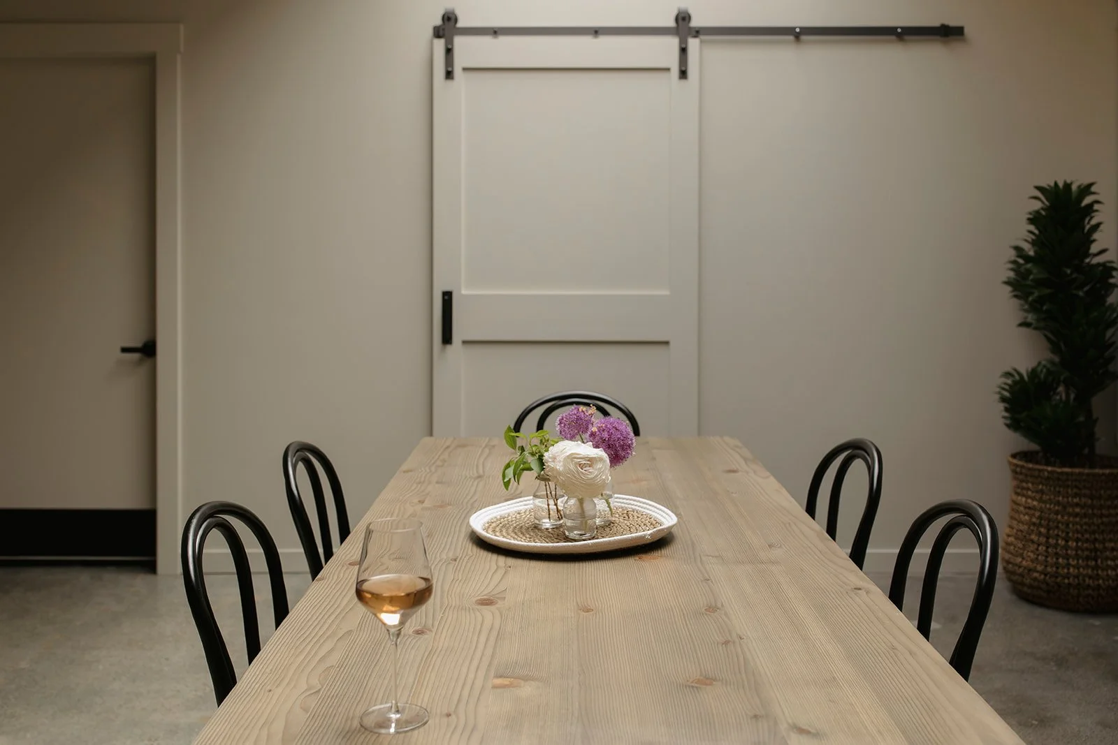 A dining room with a wooden table set with a tray holding a vase of white and purple flowers, and a glass of rosé wine. There are black chairs around the table, a large potted plant in a woven basket, a white sliding barn door, and a white interior d