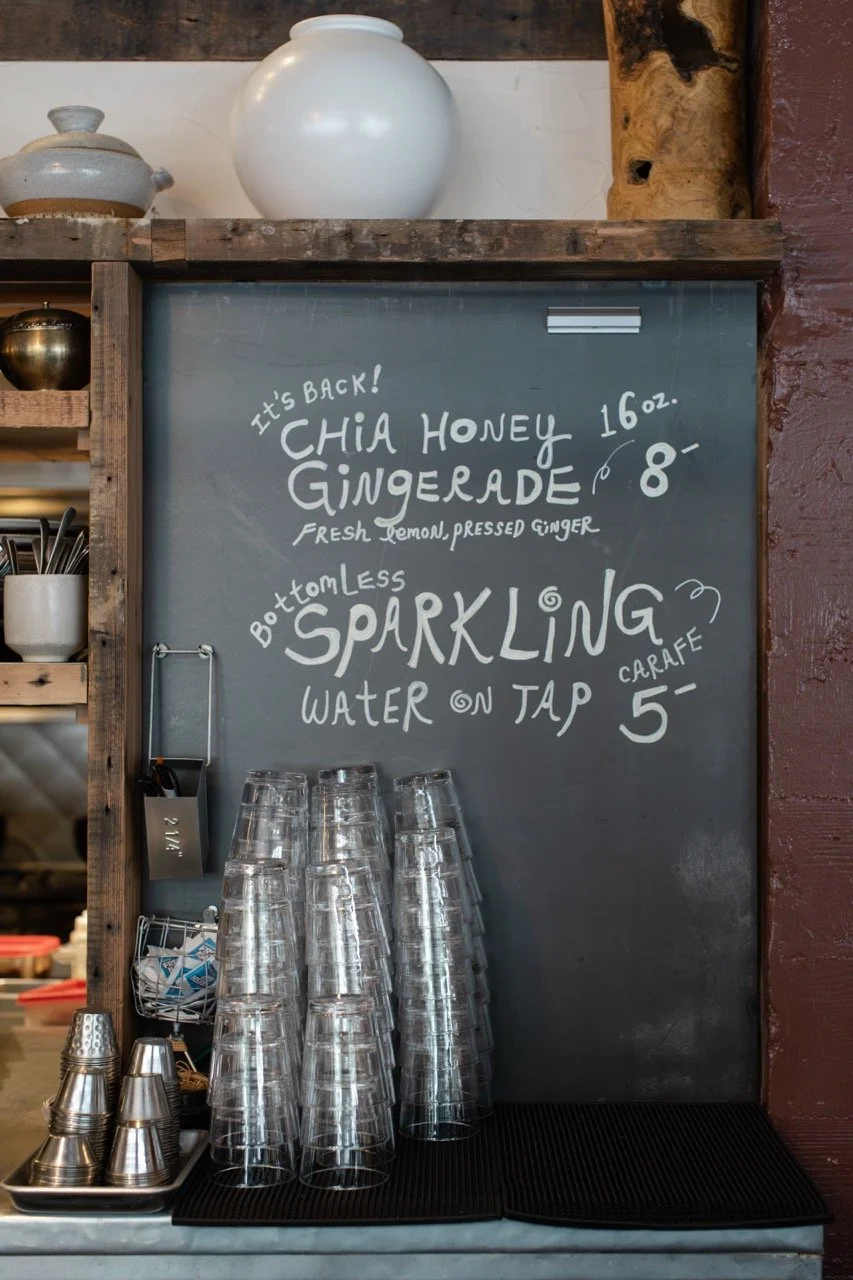 A chalkboard menu displaying prices for chia honey gingerade, sparkling water, and carafe, with stacked clear plastic glasses and metal cups below.
