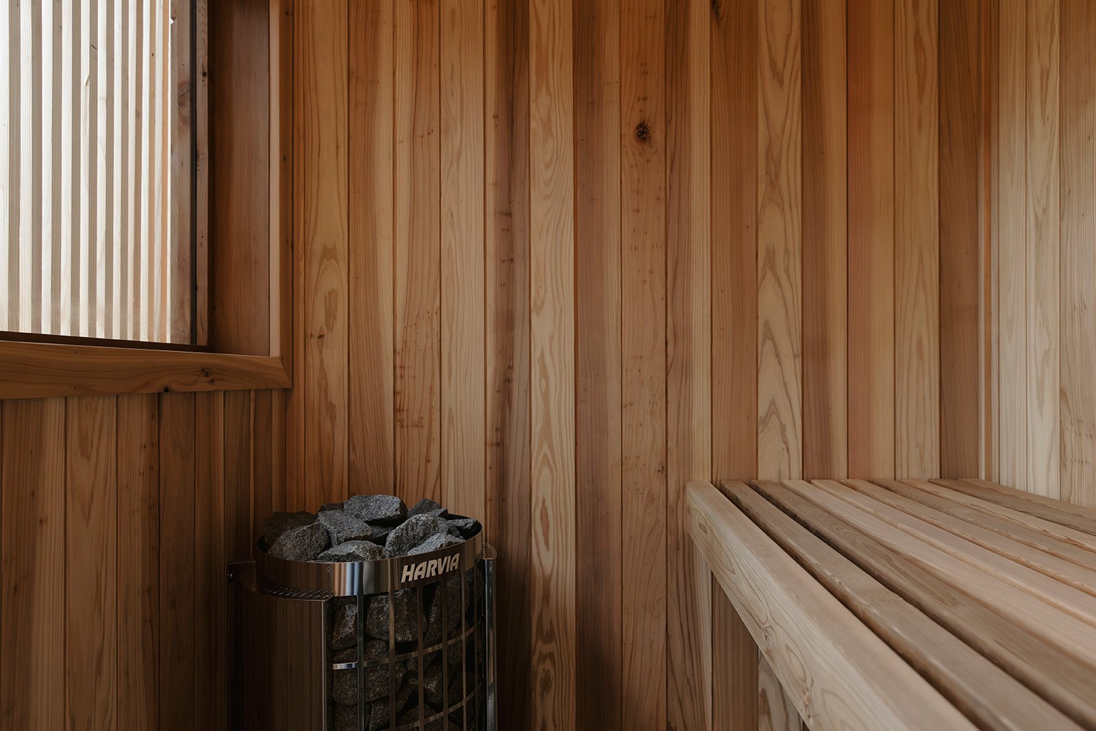 Wood-paneled interior of a sauna with a window, a wooden bench, and a heater filled with rocks.