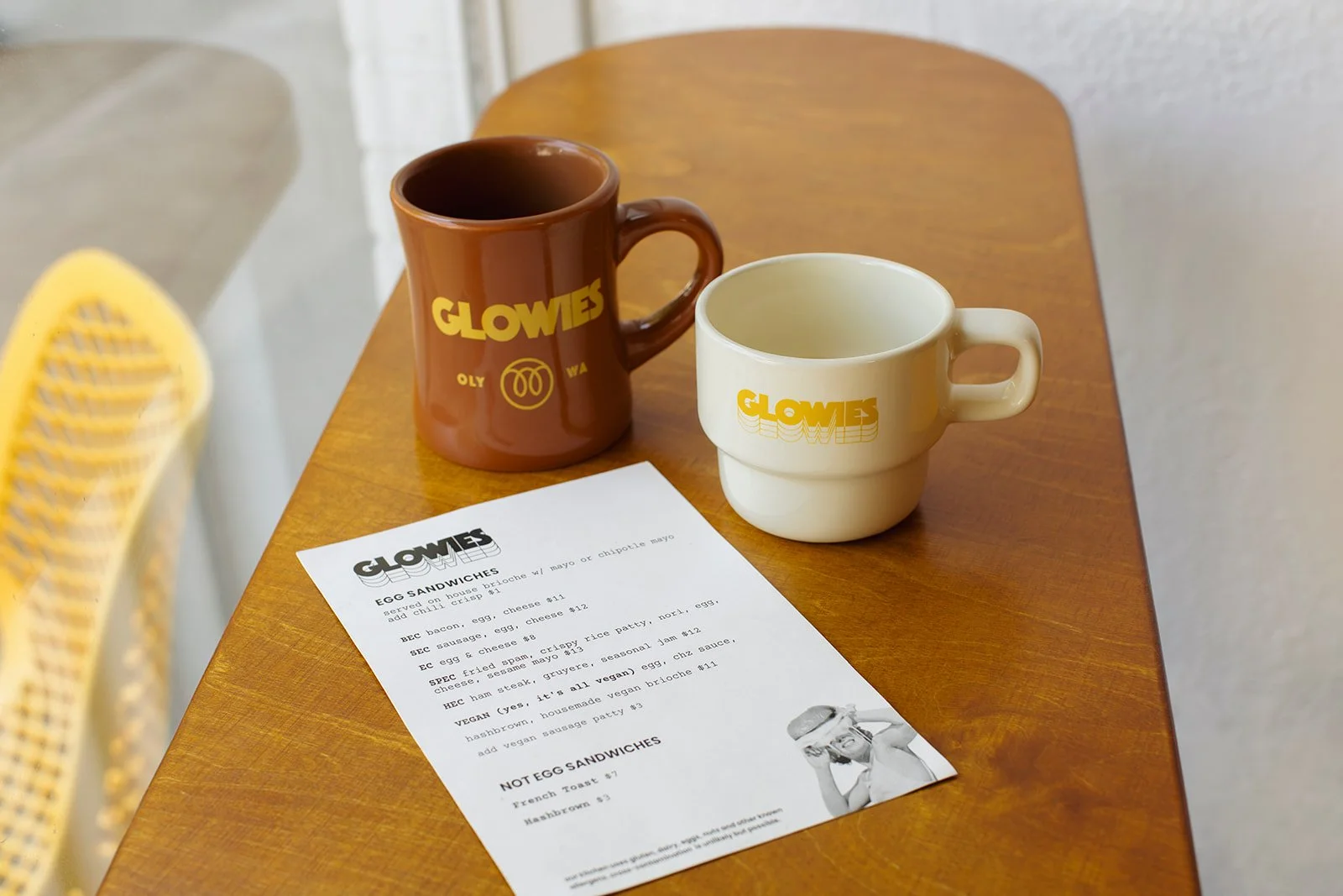 Two coffee mugs, one brown and one white, with 'GLOWES' written in yellow on both, placed on a wooden table alongside a printed menu. The background shows part of a yellow chair and a textured wall.