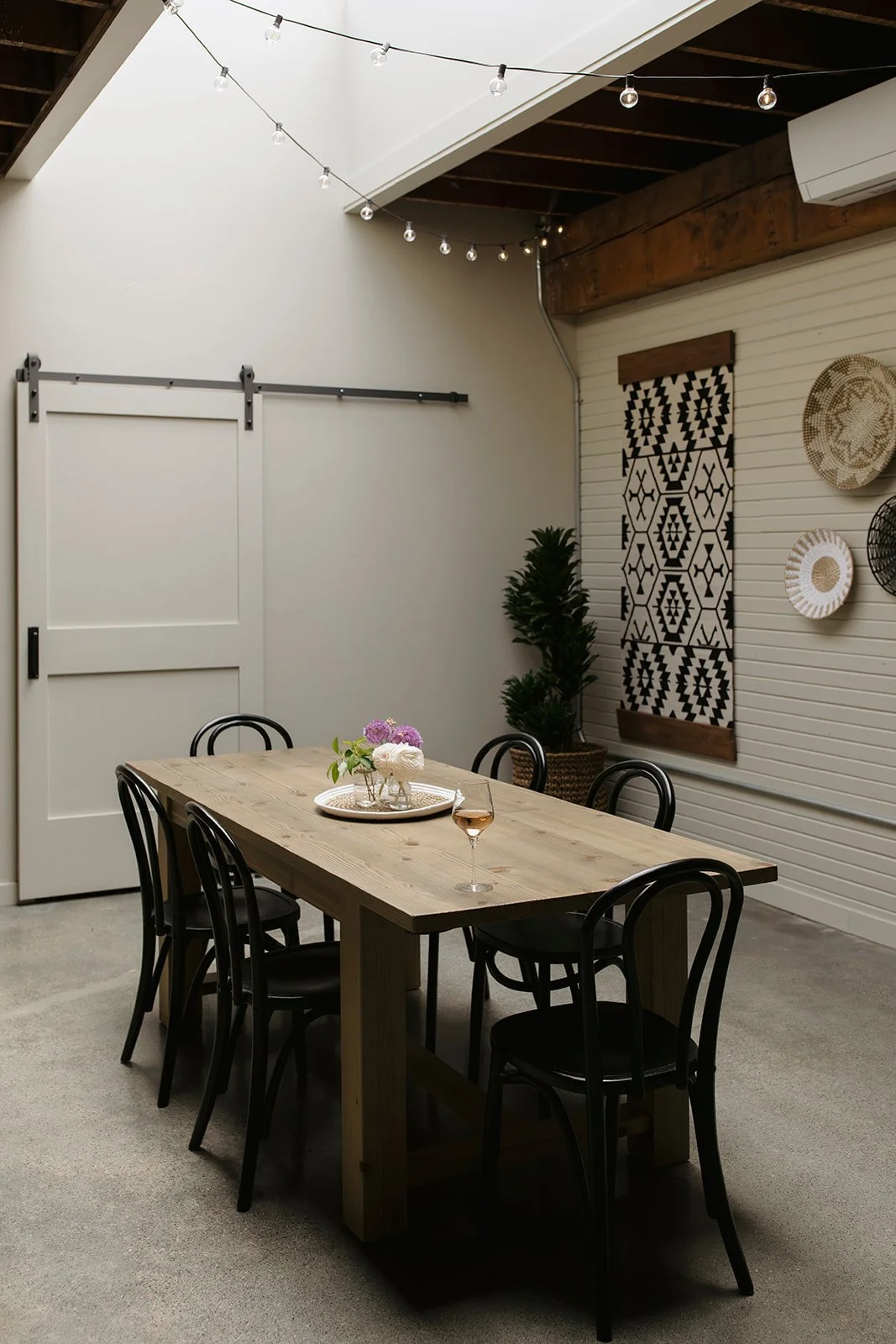 Interior of a modern dining room with a wooden table, six black chairs, a vase with purple and white flowers, a glass of rosé wine, a decorative wall hanging, wall baskets, potted plants, string lights, a barn-style sliding door, white walls, and exp