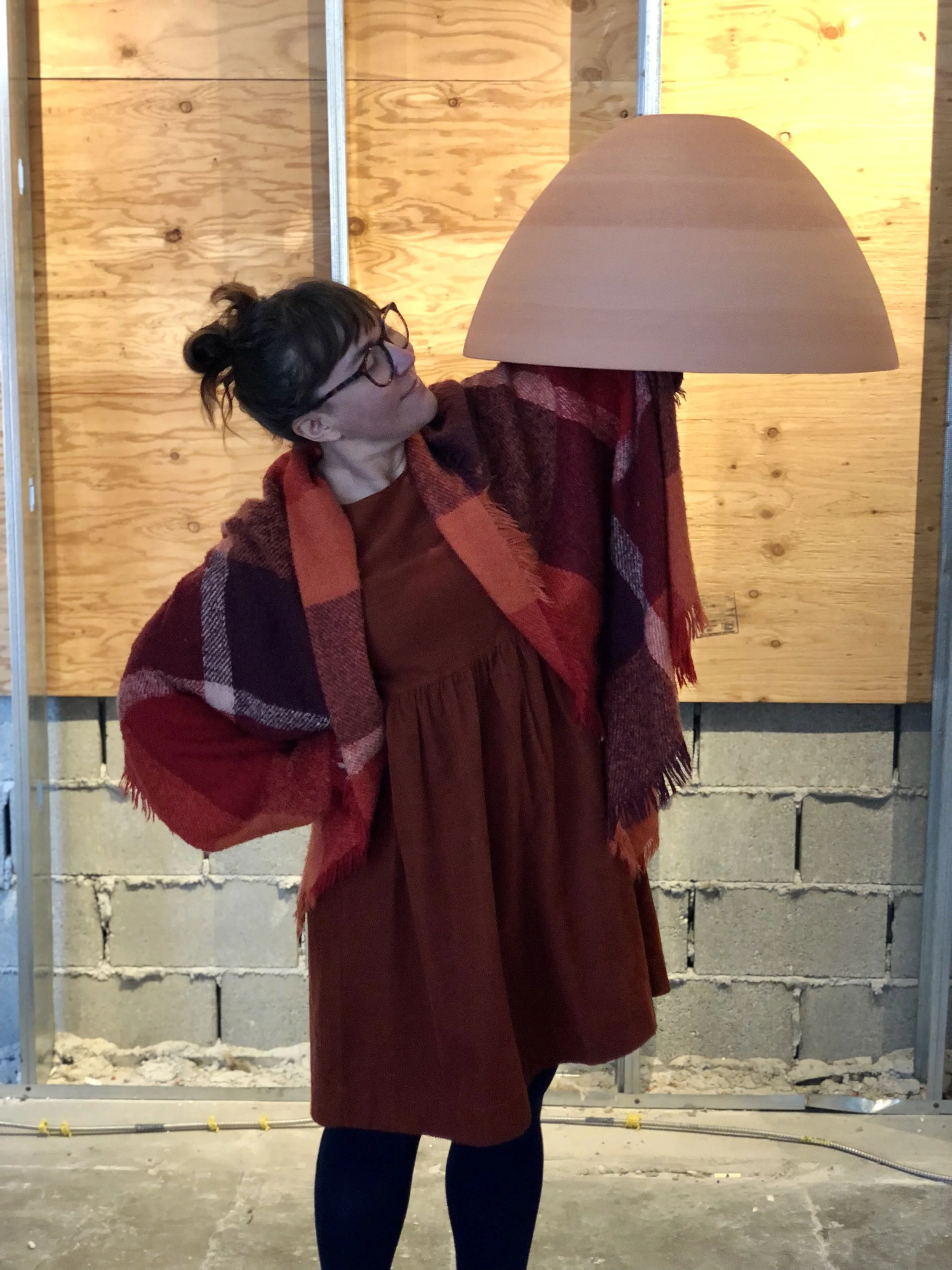 A woman wearing glasses, a brown dress, and a plaid red, brown, and beige coat, standing in front of a construction wall holding a large beige lampshade over her head.